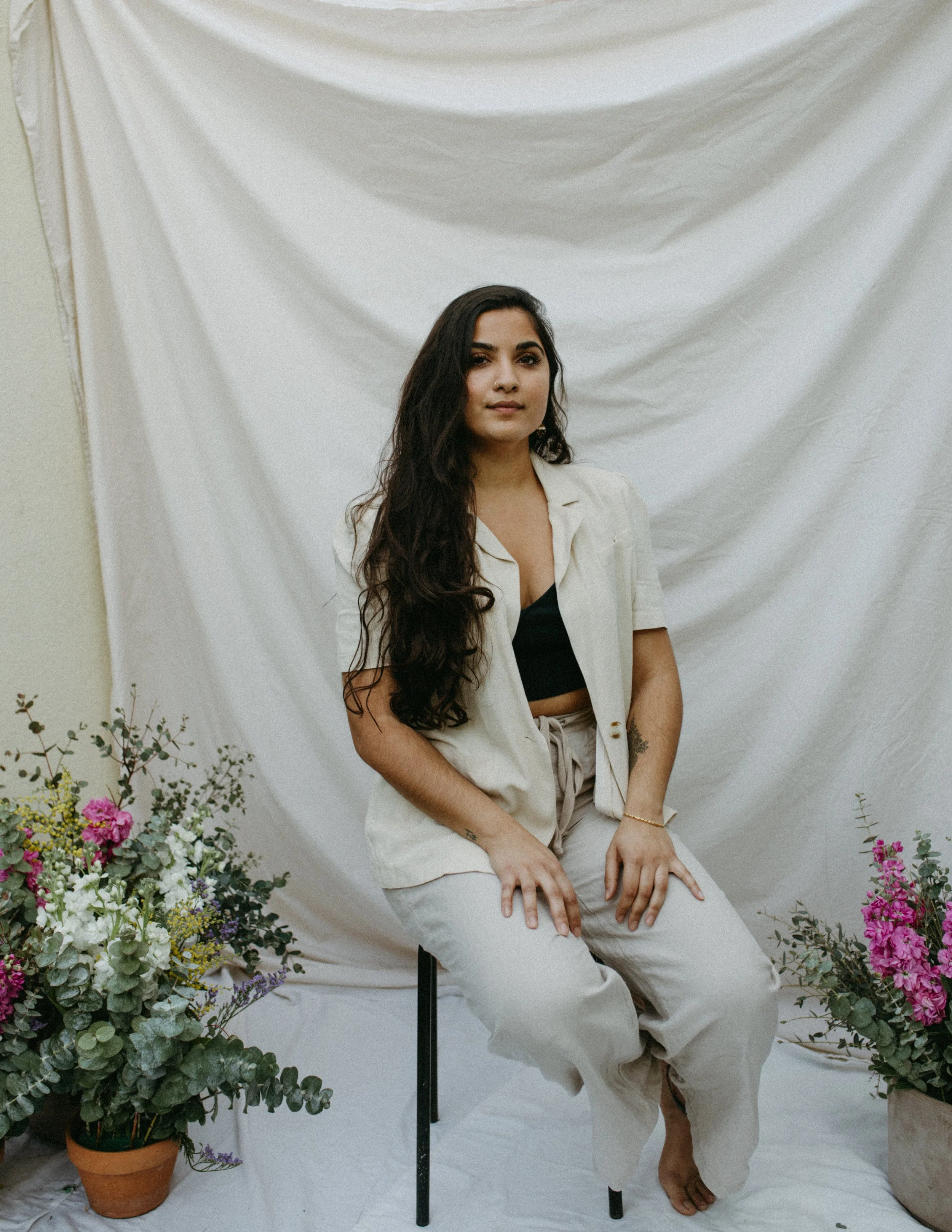 A woman with long dark wavy hair sitting on a black chair, surrounded by potted flowers, in front of a white fabric backdrop.