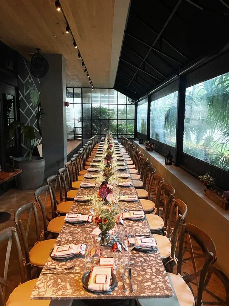 Long rectangular dining table set for a formal event with floral centerpieces, arranged in a bright room with large windows and natural light, with chairs on both sides.