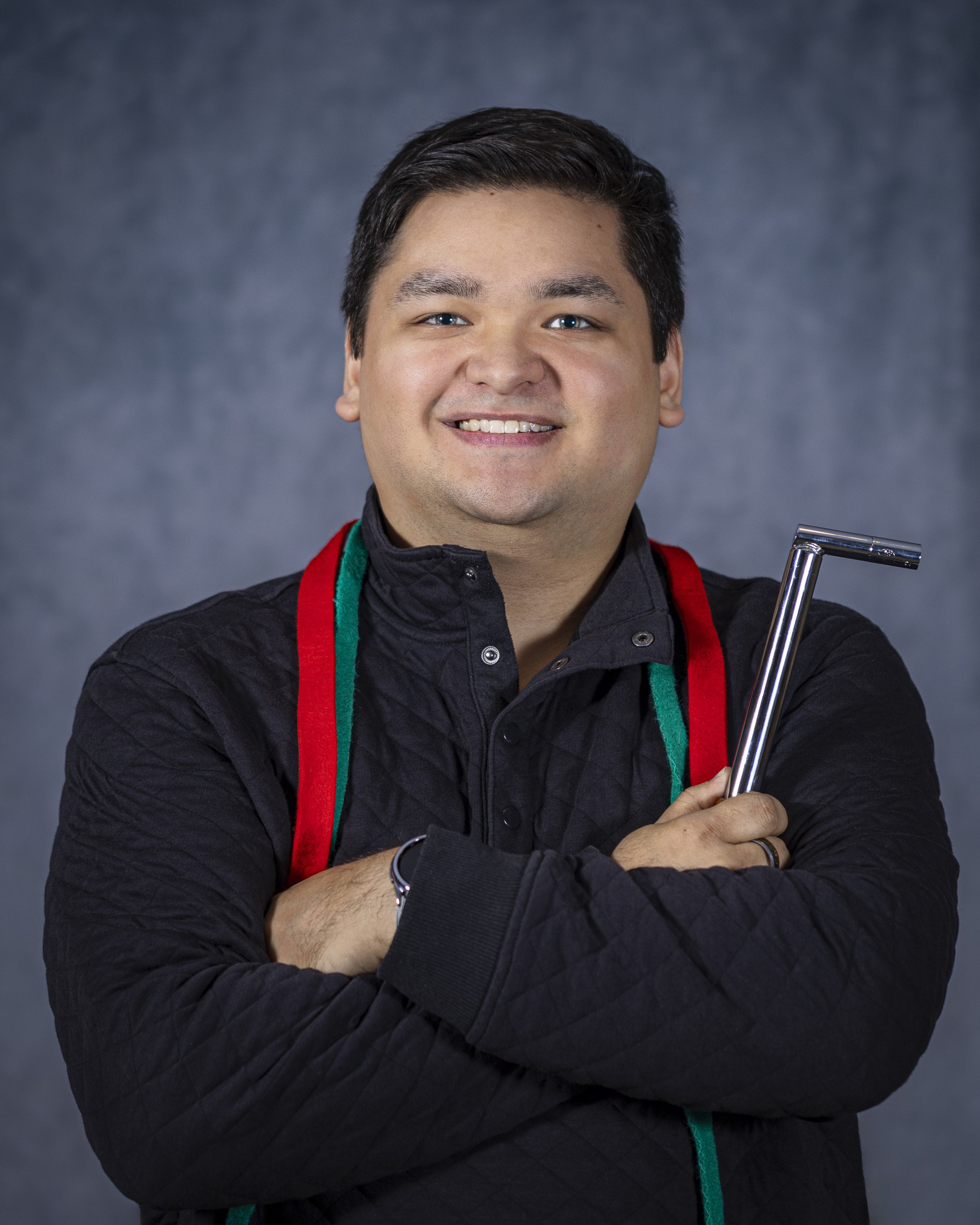 Piano tuner holding a tuning hammer tool, wearing a black jacket and red and green felt strips made for piano tuning, posing against a blue background.