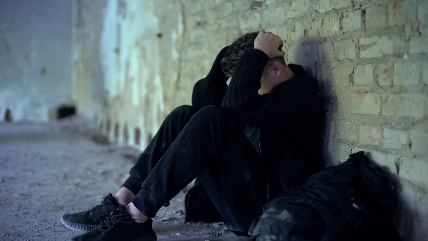 Young man sitting on the ground with his back against a brick wall, holding his head in his hands, appearing distressed, in an abandoned setting.