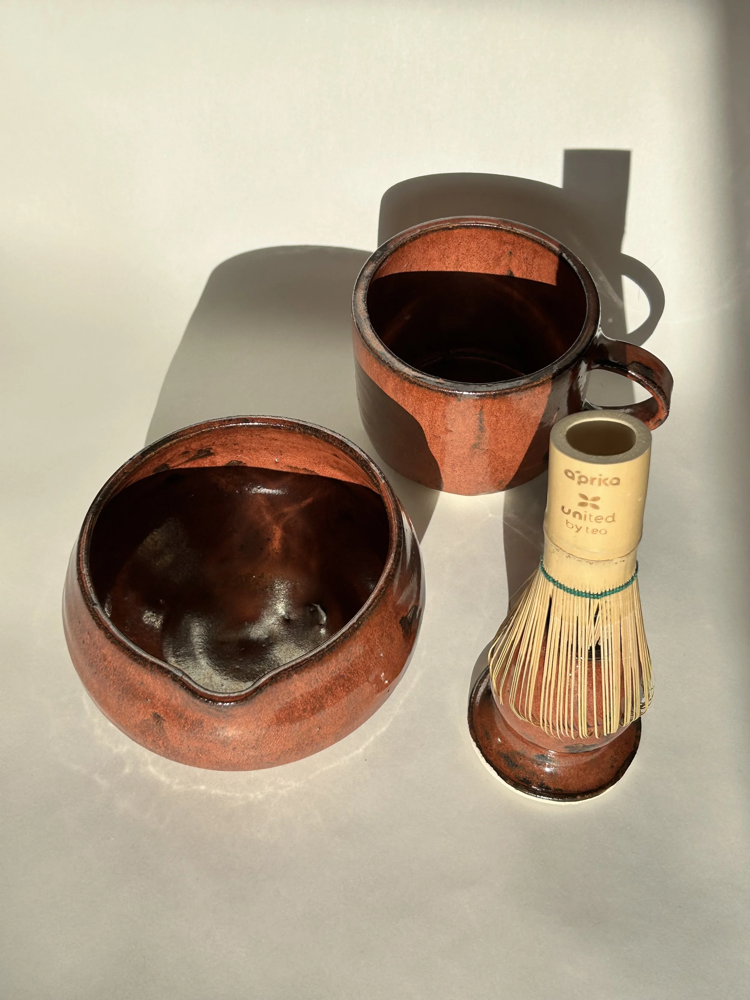 Two brown ceramic cups with dark liquid and a small ceramic container with a bamboo whisk, used for preparing matcha tea, on a white surface with shadows.