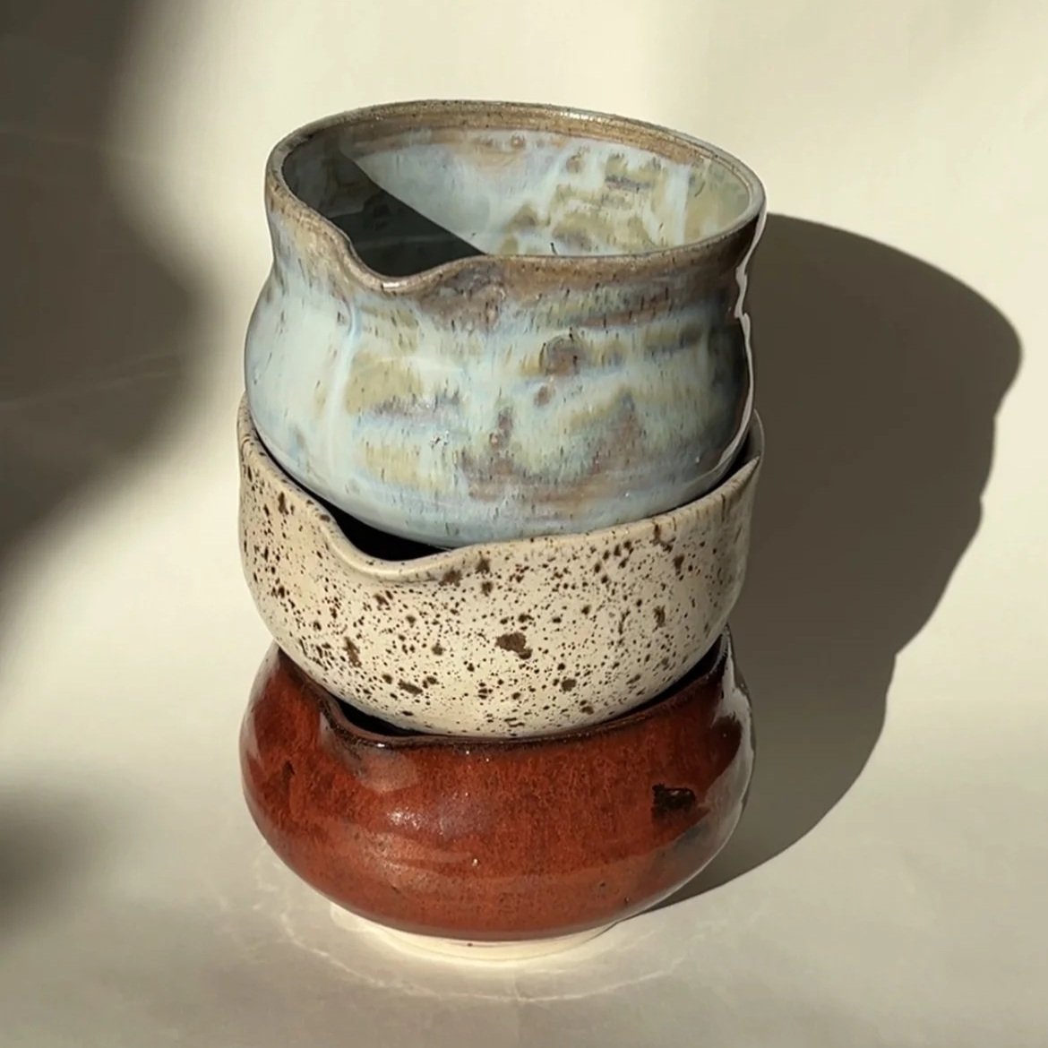 A stack of four ceramic bowls with different glaze finishes, cast against a light background with a shadow.