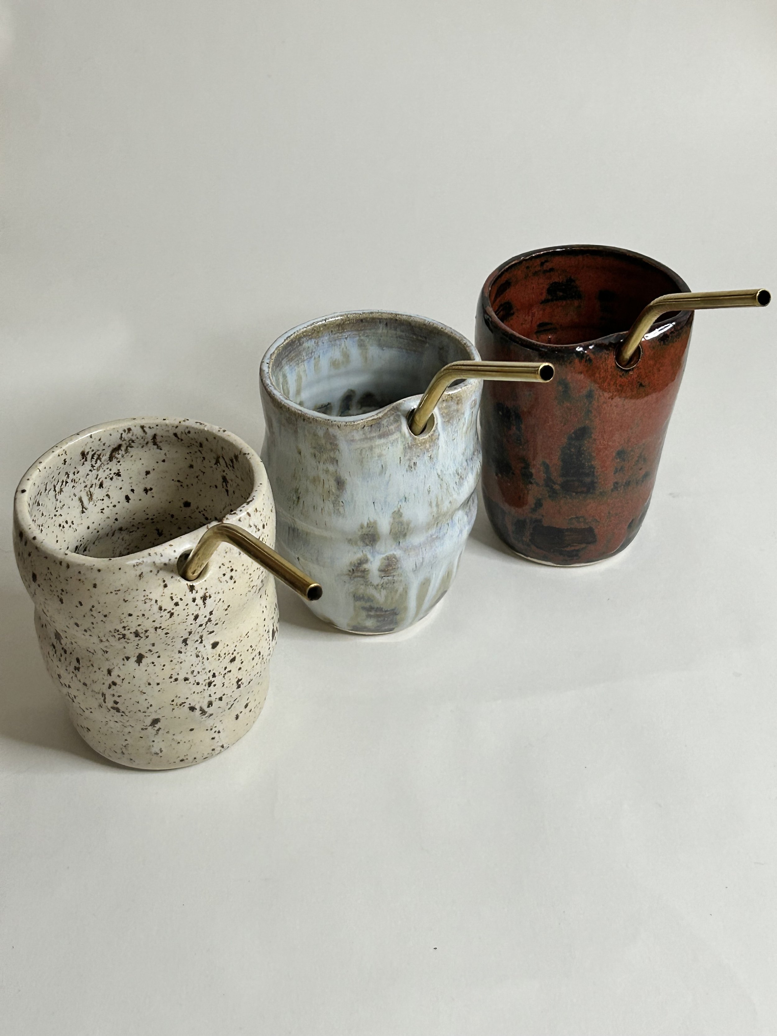 Three ceramic tumblers with metallic straws, each with different glaze designs in beige, blue, and red, placed on a white surface.