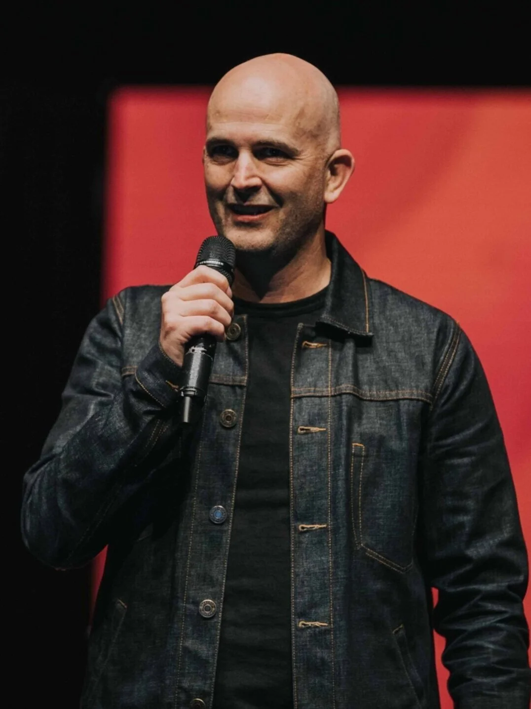 A bald man in a black denim jacket holding a microphone speaks at an event against a red background.