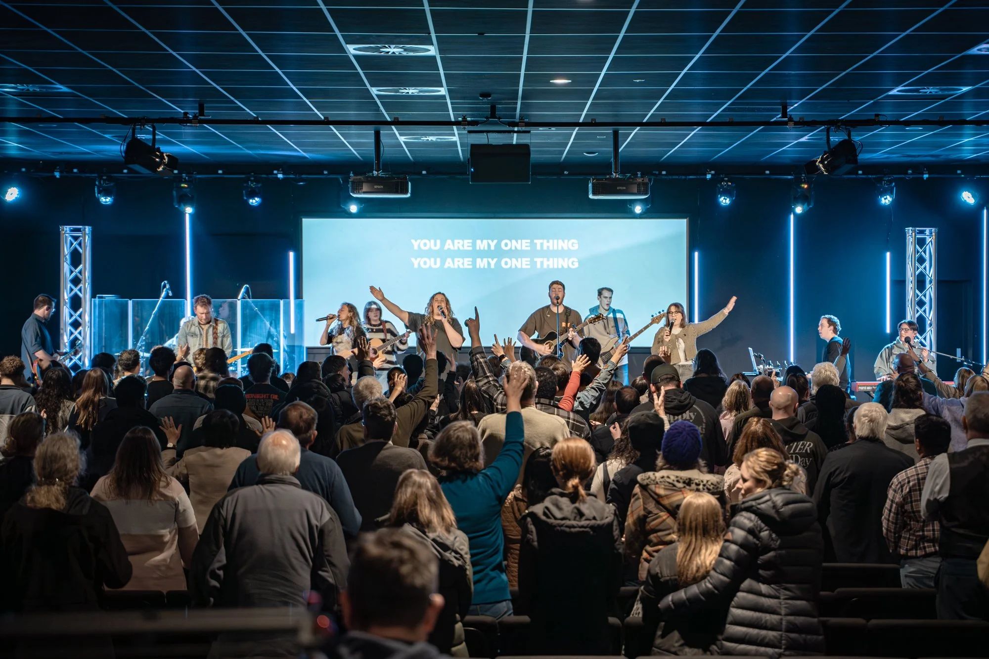 A band performs on stage with a large screen behind them displaying the lyrics, 'You are my one thing,' while a large audience listens, raises their hands, and engages in the music.