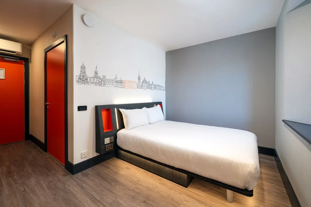 A modern hotel room with a white bed, black and red headboard, and city skyline mural on the wall. The room has wood flooring, gray and white walls, a window with a sill, and red doors, one leading to a closet and one likely to a bathroom.