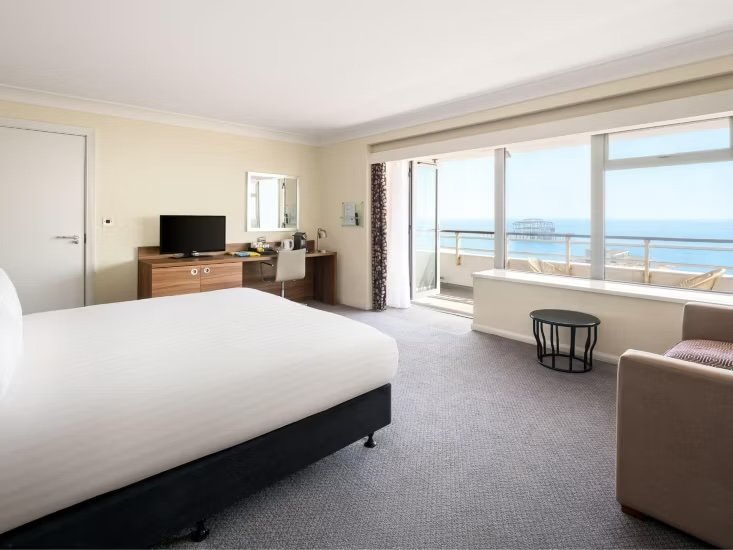 Hotel room with a large bed, a television on a wooden dresser, a desk with a mirror and chair, sliding glass doors opening to a balcony with ocean view, and a small sitting area with an armchair and side table.