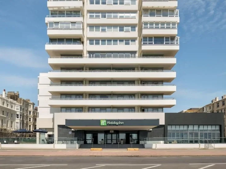 Front view of a tall white multi-story building with balconies, housing a Holiday Inn hotel on the ground floor.