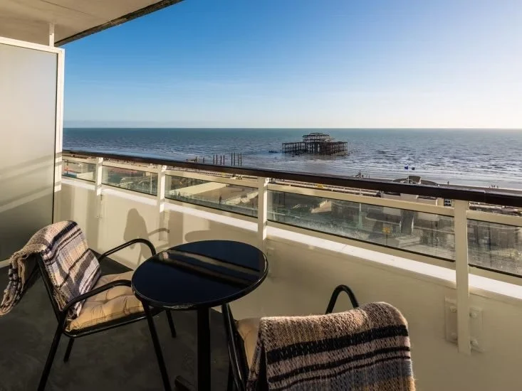 Balcony with two chairs with blankets, a small round table, overlooking a beach with a pier extending into the ocean under a clear blue sky.