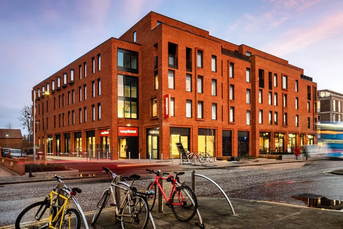 Modern red brick hotel building with large windows, bicycles parked outside, and a street with moving vehicles at dusk.
