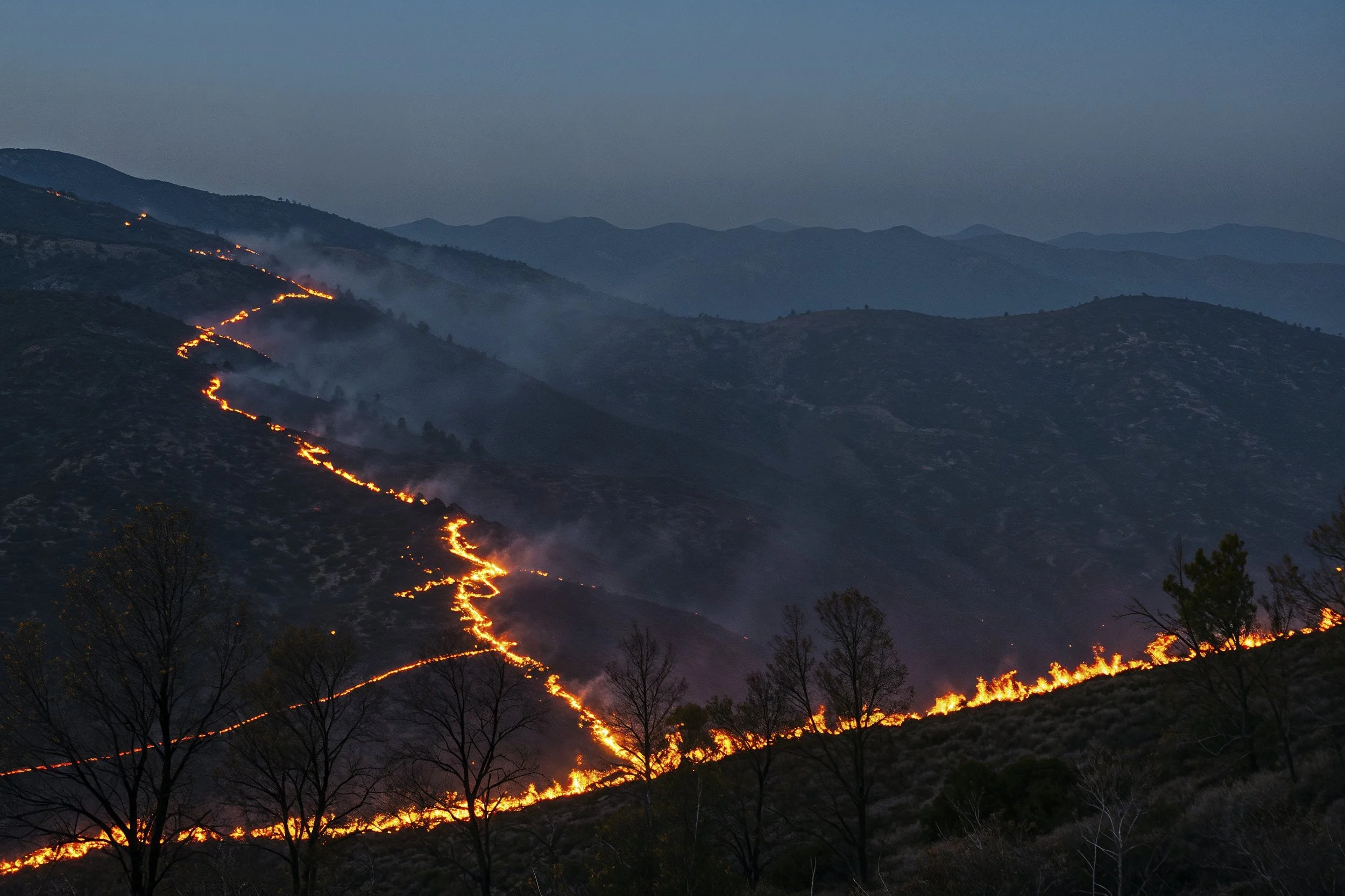 A wildfire burning along the mountain ridge, with flames visible on the hillside and smoke rising into the sky at dusk.