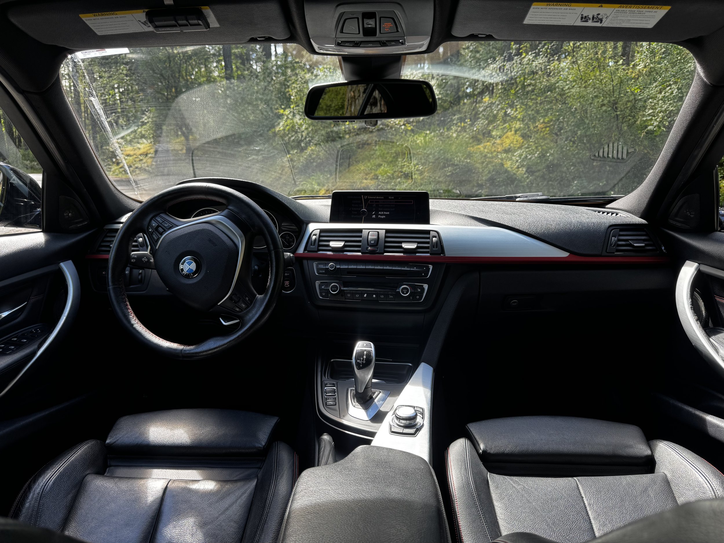 Inside view of a BMW vehicle's dashboard and front seats, showing the steering wheel, center console, gear shifter, and an infotainment screen with trees visible through the windshield.