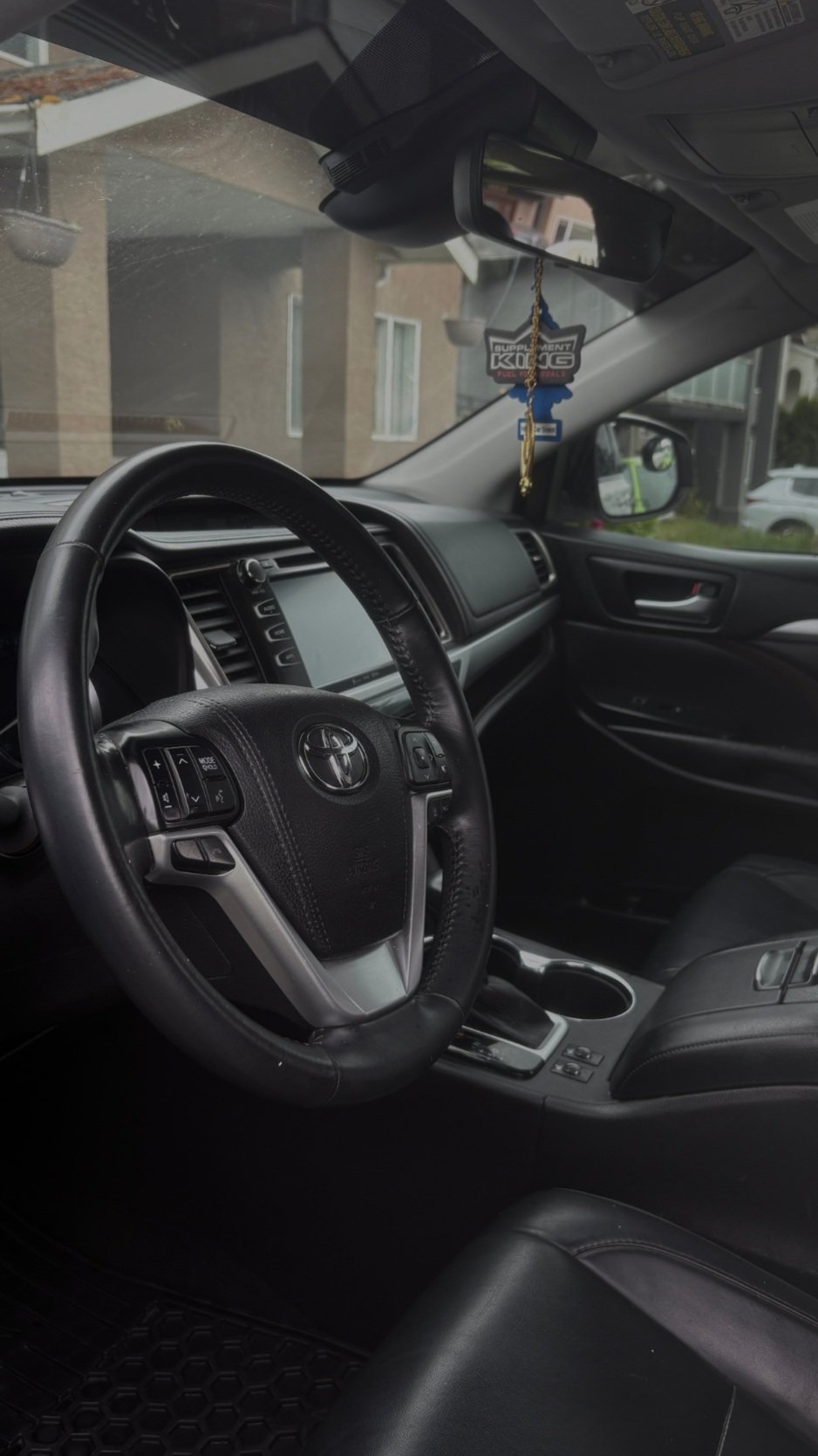 Interior of a Toyota vehicle showing the steering wheel, dashboard, and front seats, with rearview mirror and house visible through the windshield.