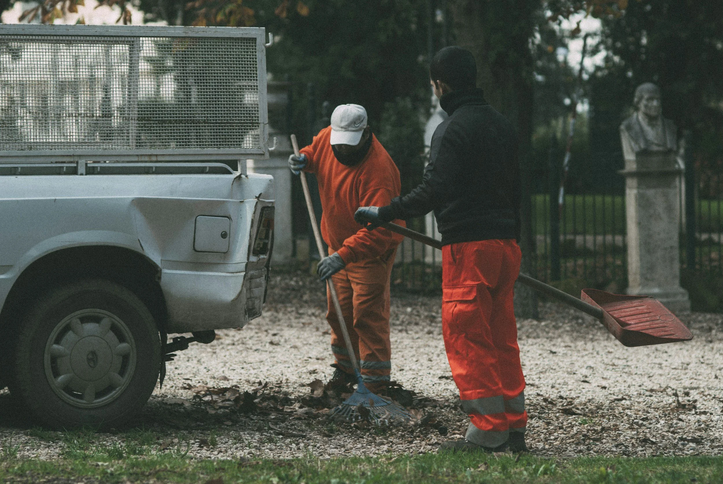 Two workers in orange and black clothing and gloves are cleaning up fallen leaves and debris near a white truck with a metal cage on its bed, in an outdoor park area with trees and bust sculptures in the background.