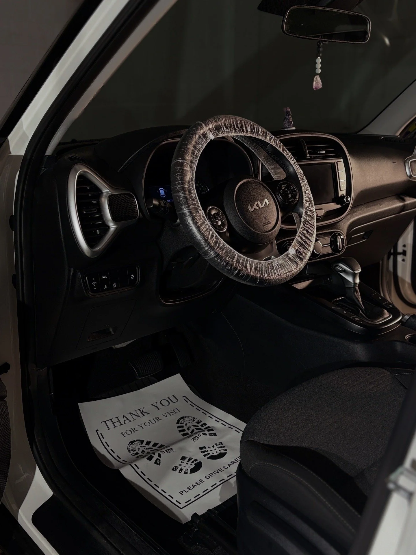 Interior of a Kia car with a plastic-wrapped steering wheel, dashboard, gear shift, and a paper floor mat that says "Thank You for Your Visit" with footprints, placed on the driver's side floor.
