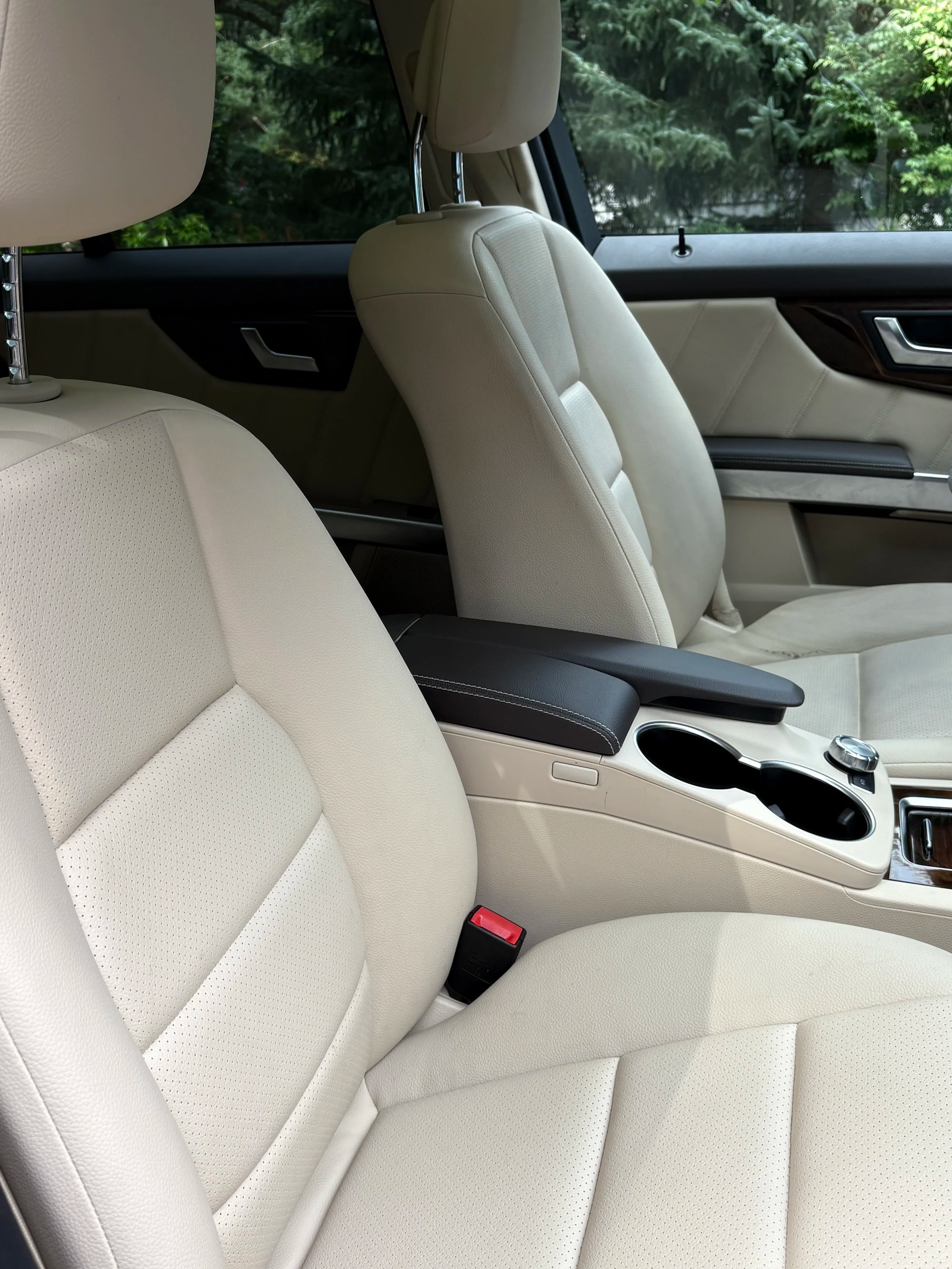 Interior of a luxury car with cream-colored leather seats, a center console with cup holders, and a view of green trees through the window.