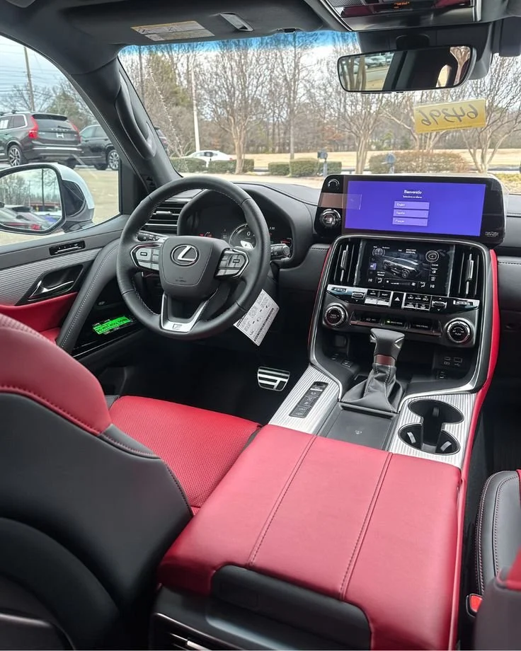 Interior of a Lexus vehicle, showing the steering wheel, dashboard, center console, and red and black leather seats, with a screen displaying a welcome message.