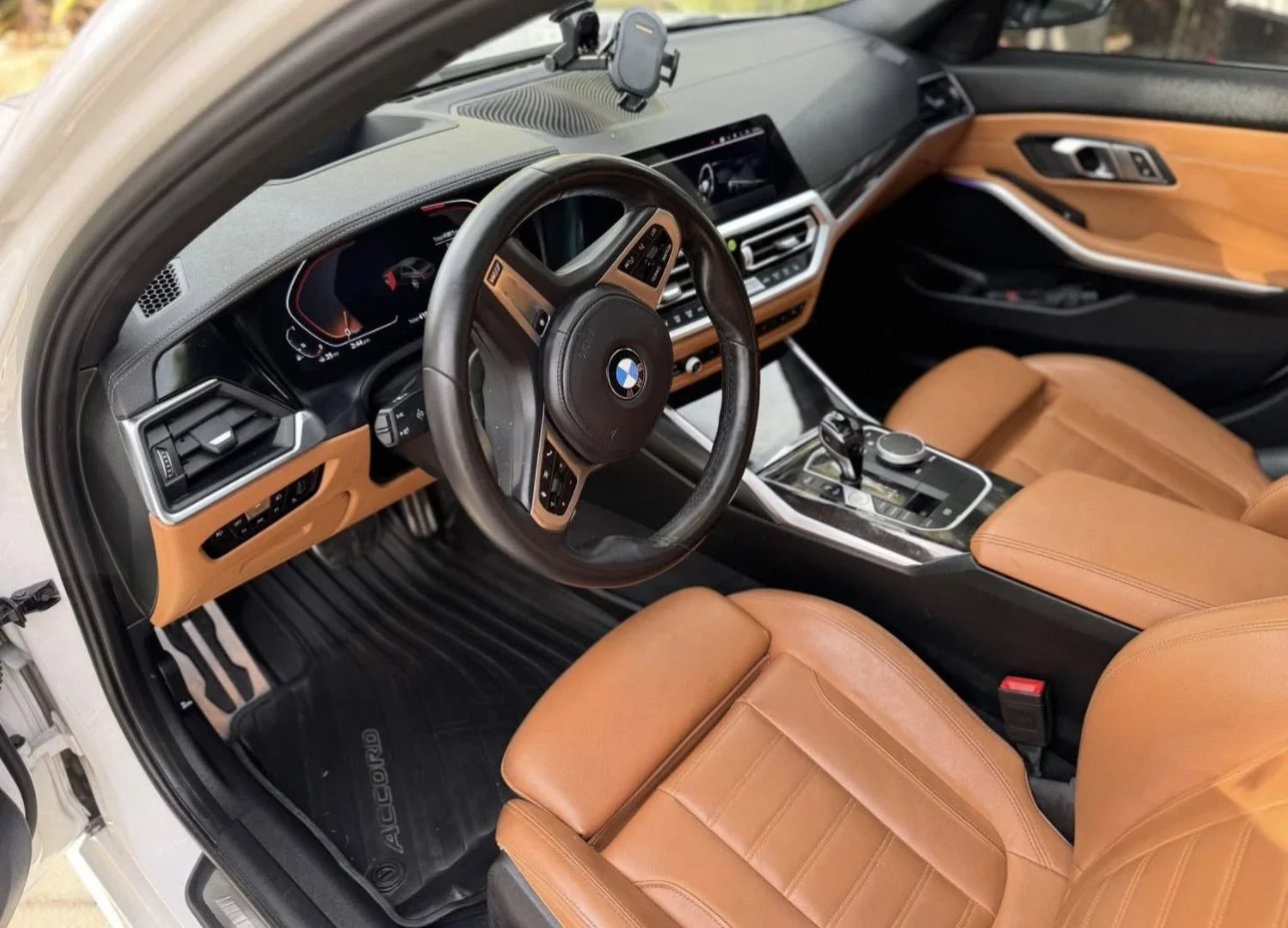 Interior of an BMW car showing a tactile dashboard, leather seats, steering wheel with BMW logo, center console with control knob, and digital instrument panel.