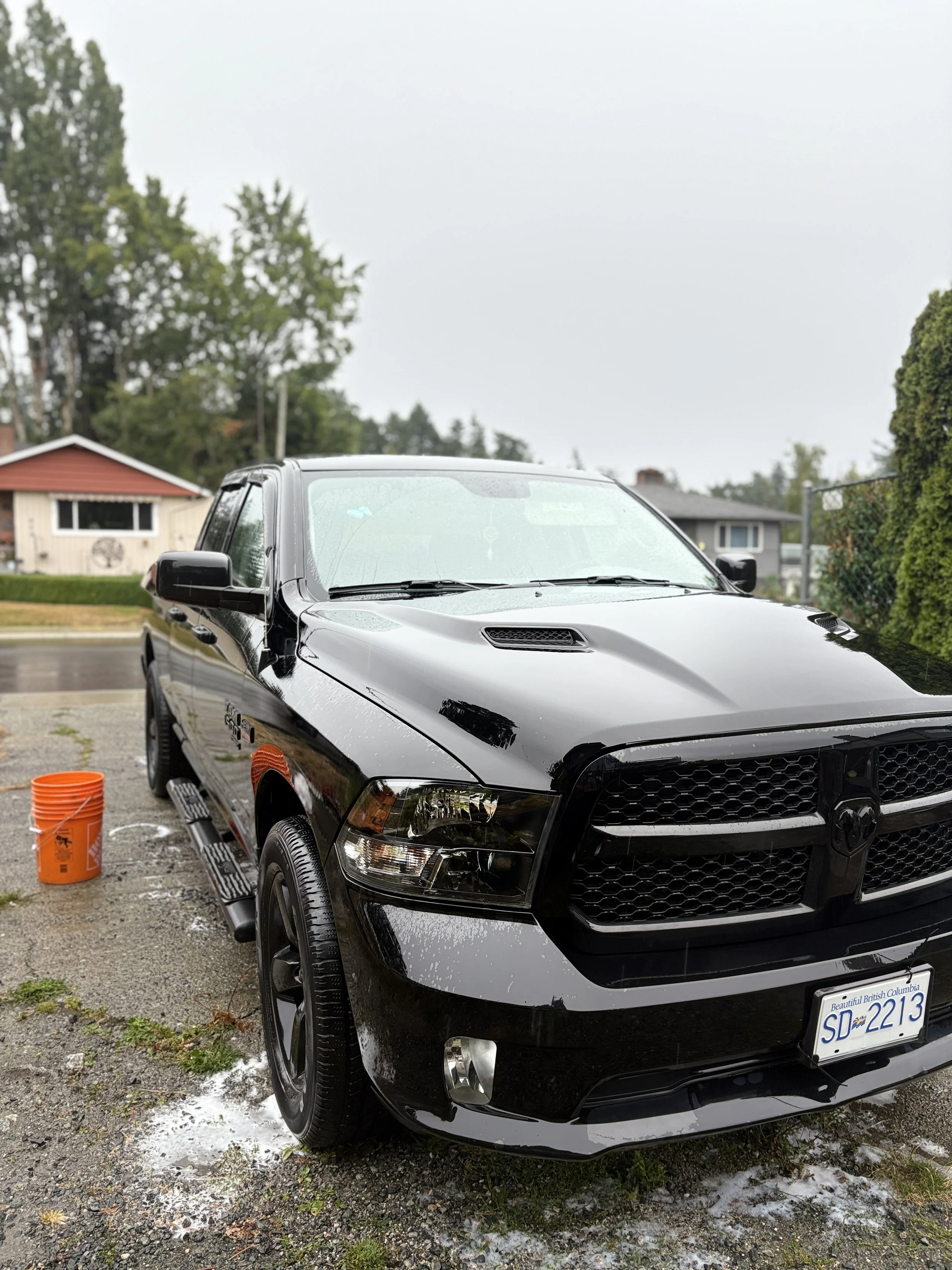 A black pickup truck is being washed and is covered with soap suds, with an orange bucket and a wet soap streak on the ground nearby, in a residential driveway on a rainy day.