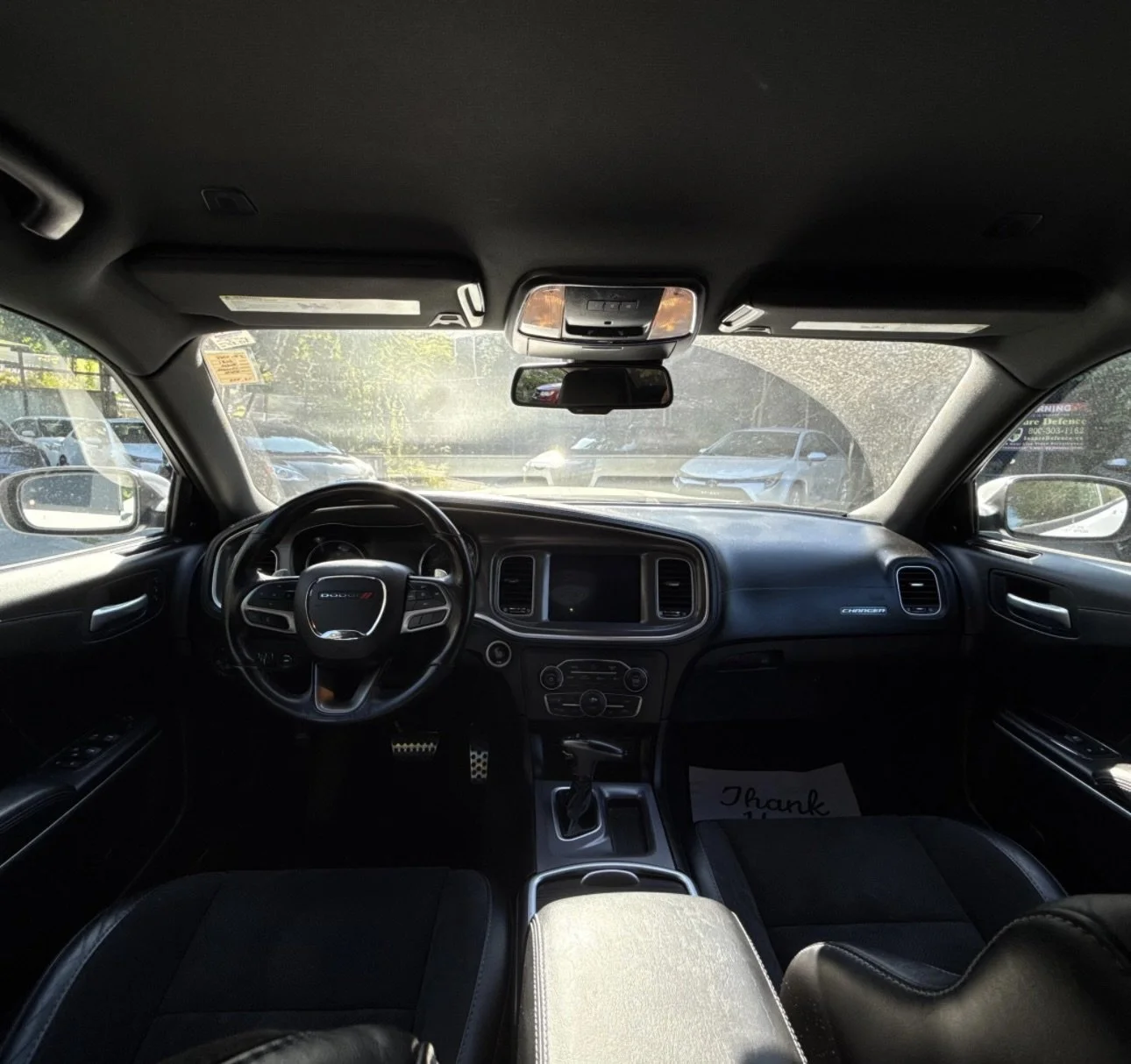 Interior view of a Dodge Charger vehicle with black dashboard, steering wheel, and front seats, parked in a lot with other cars visible through the windows.