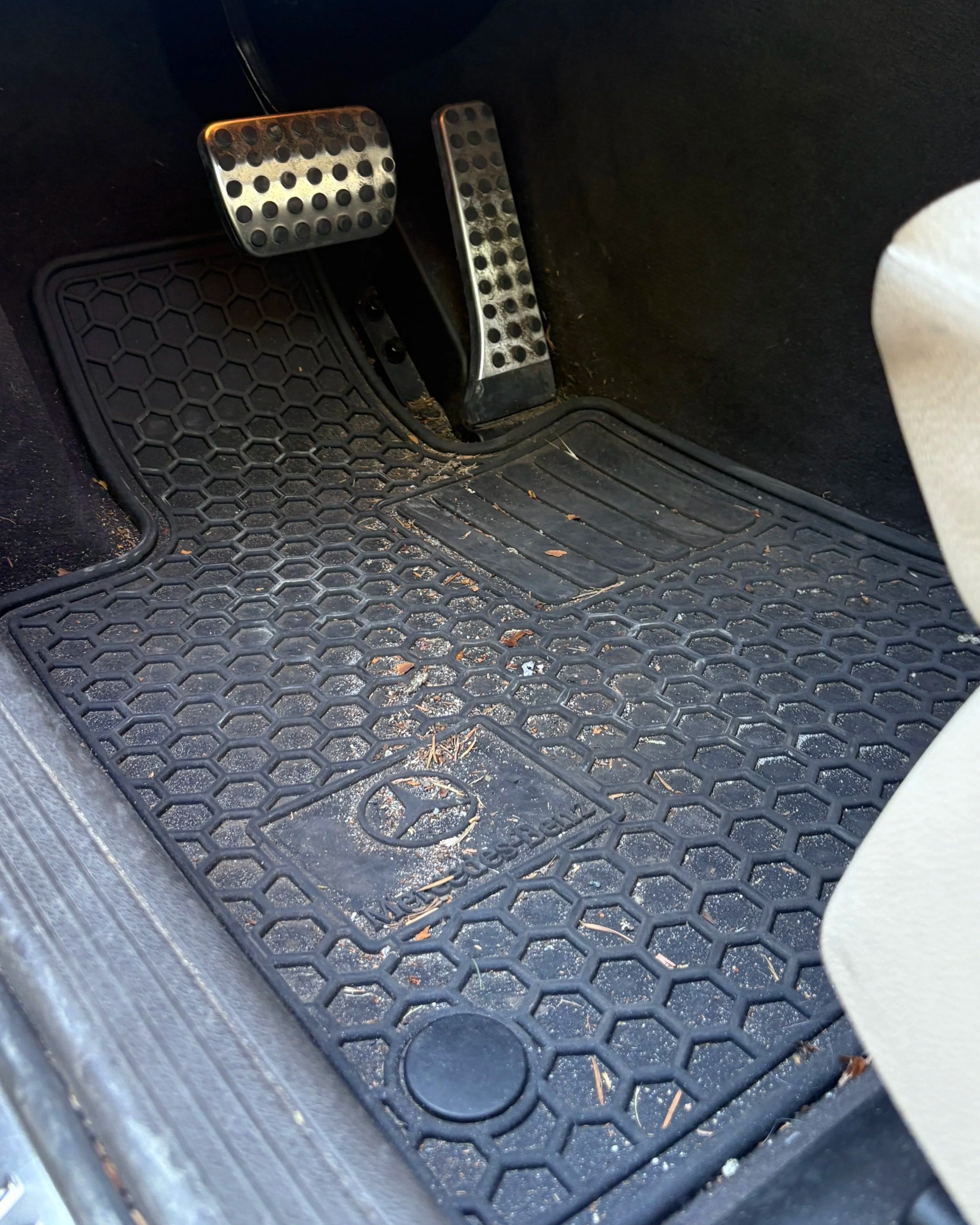 The driver side footwell of a car showing brake and accelerator pedals, with a dirty rubber floor mat.