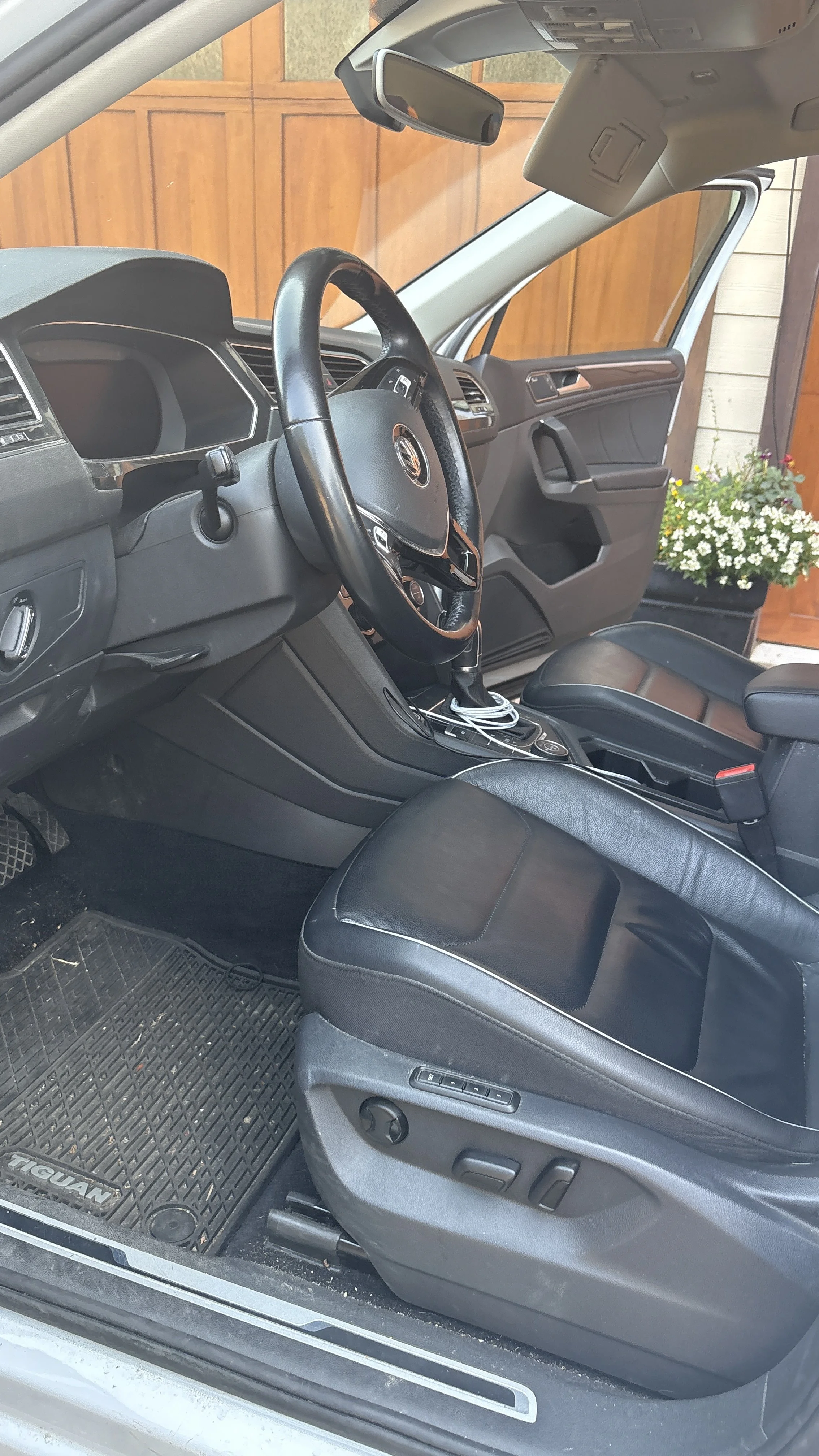 Interior of a vehicle showing the driver's seat, steering wheel, dashboard, center console with charging cable, and a passenger seat. The vehicle is parked with a wooden fence and potted flowers visible outside.