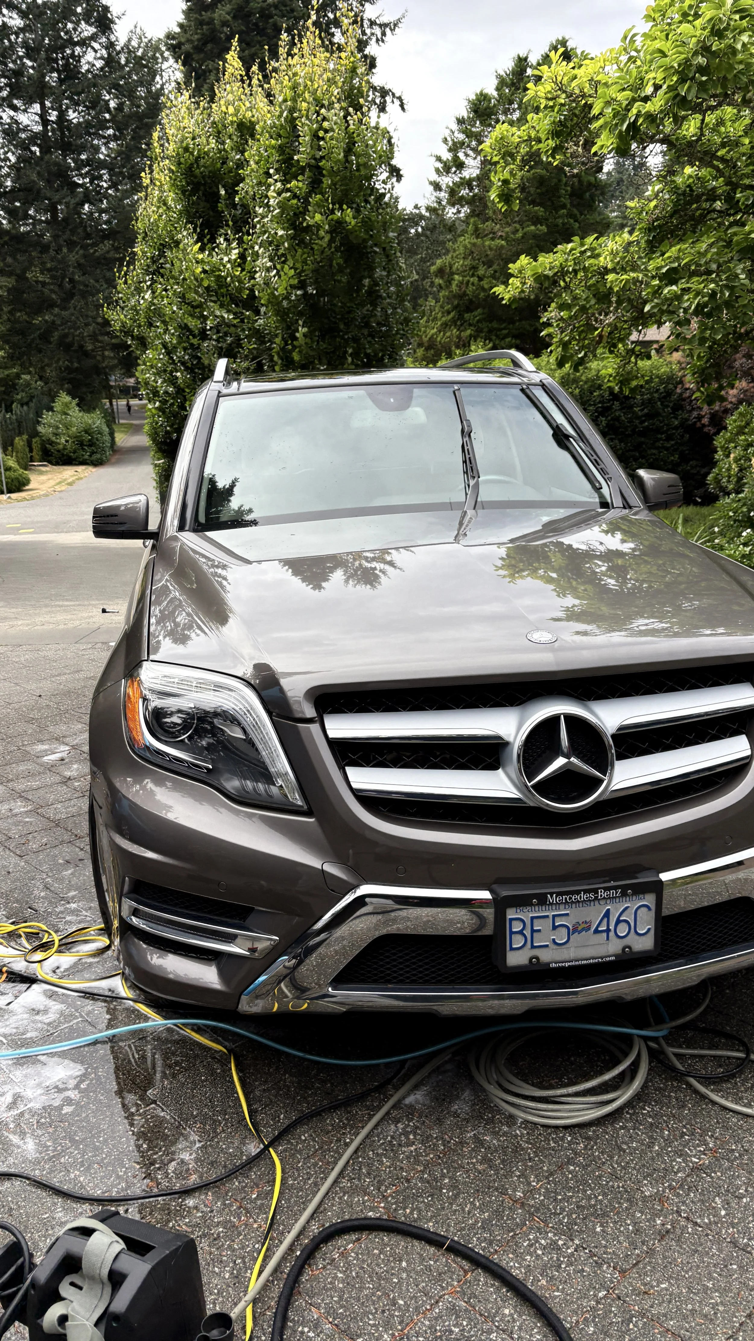 A brown Mercedes-Benz SUV parked outdoors on a driveway, connected to various electrical cables and equipment, with trees and greenery in the background.