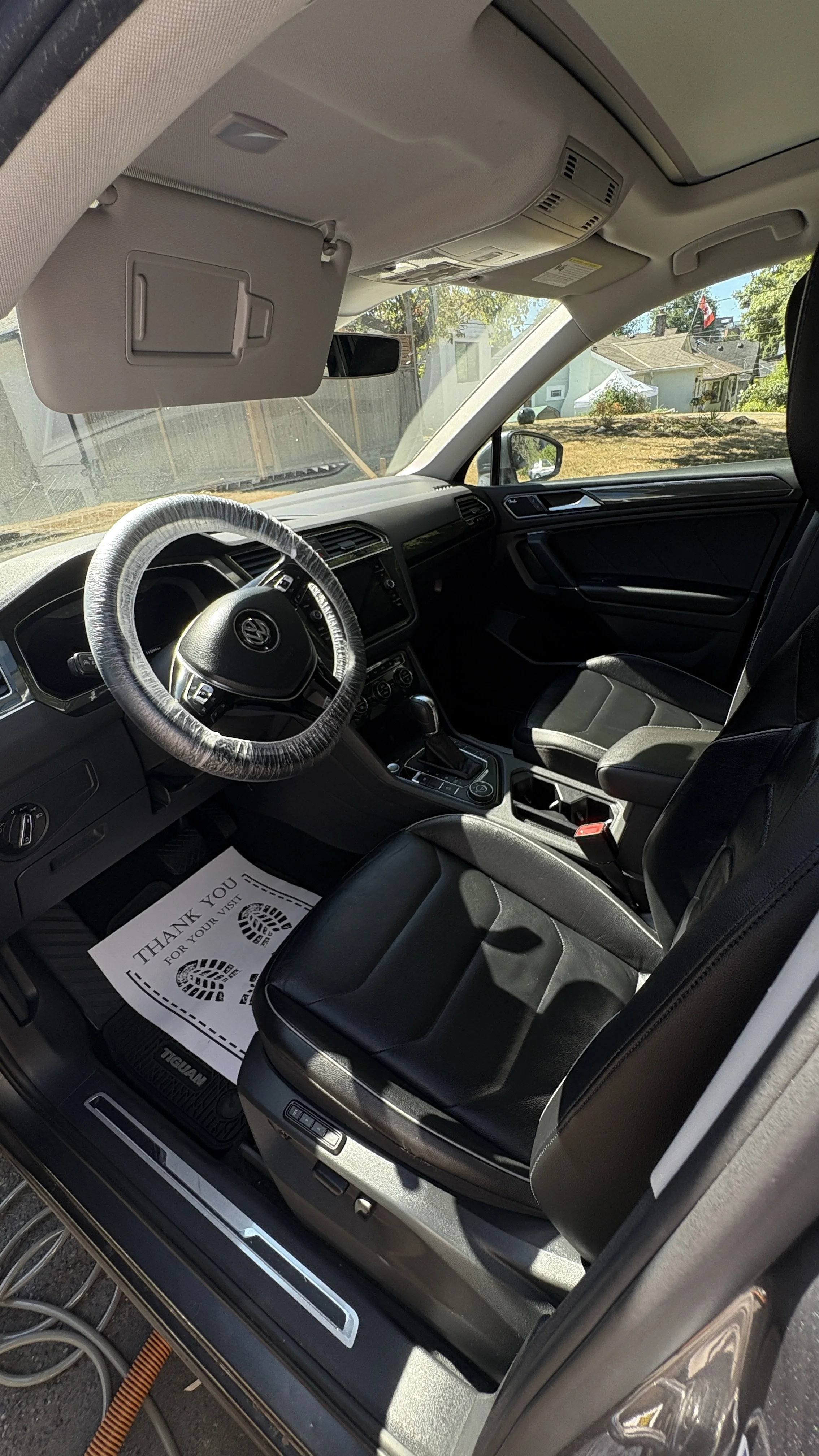 Interior of a Volkswagen Tiguan SUV showing the driver's seat with a plastic cover on the steering wheel, center console, dash, and a paper floor mat reading "Thank you for your visit". Outside, some houses and trees are visible through the windshield and window.