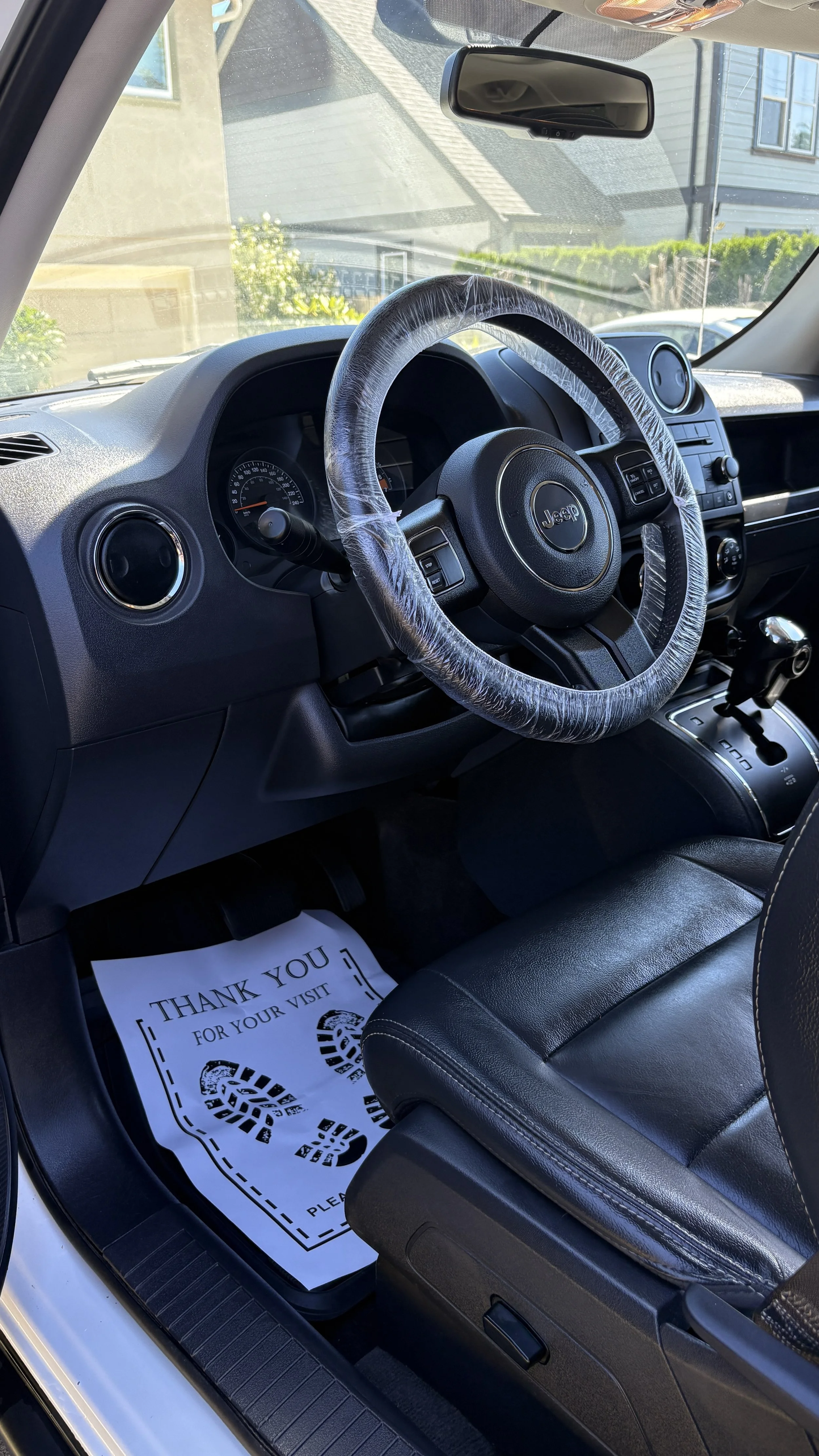 The interior of a Jeep vehicle showing the steering wheel with a plastic cover, black leather seat, and a paper floor mat that reads 'Thank you for your visit.'