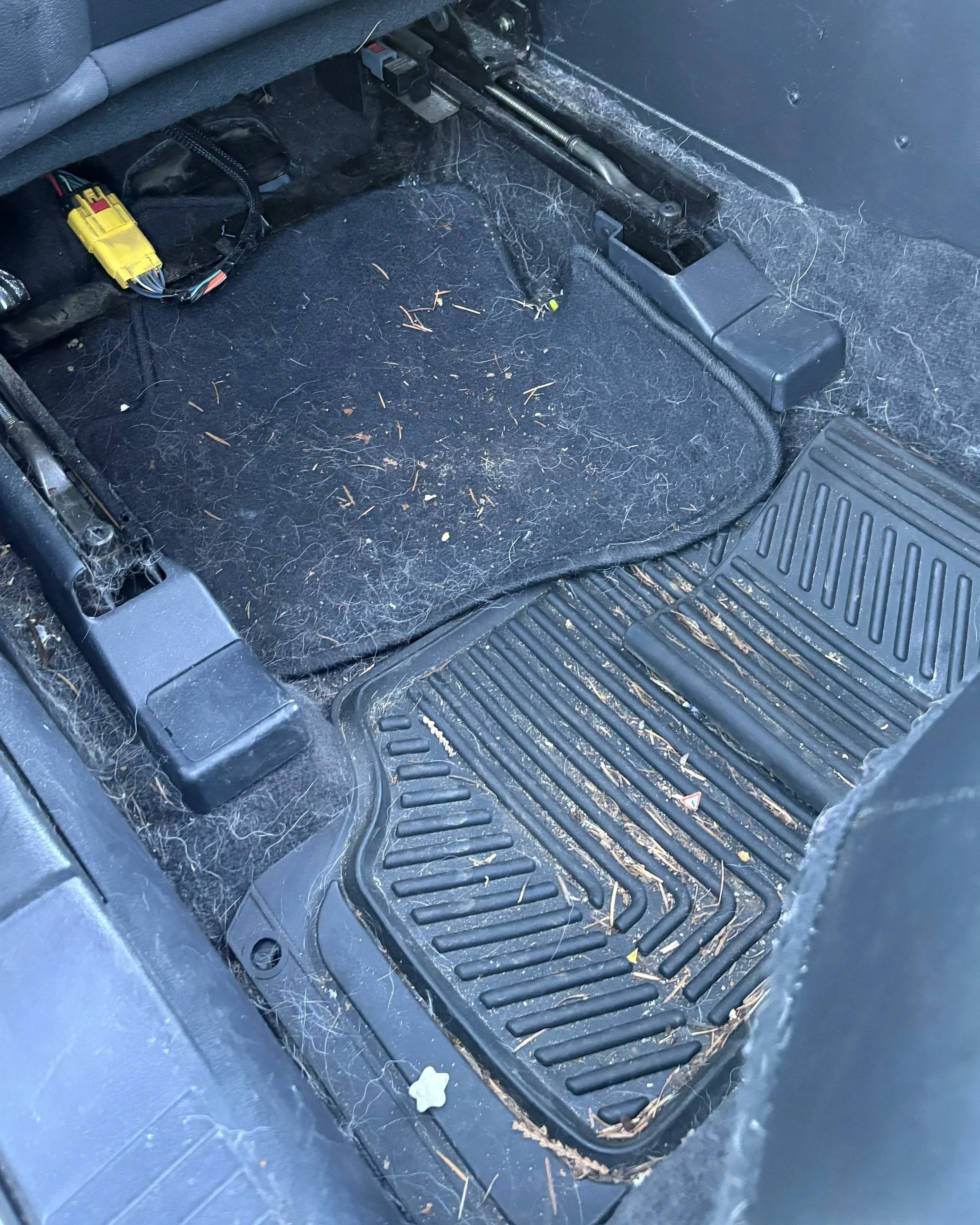 Car floor mat covered in dust, hair, dirt, and small debris, with visible dirt stains and a yellow wire connector connected to a wiring harness.