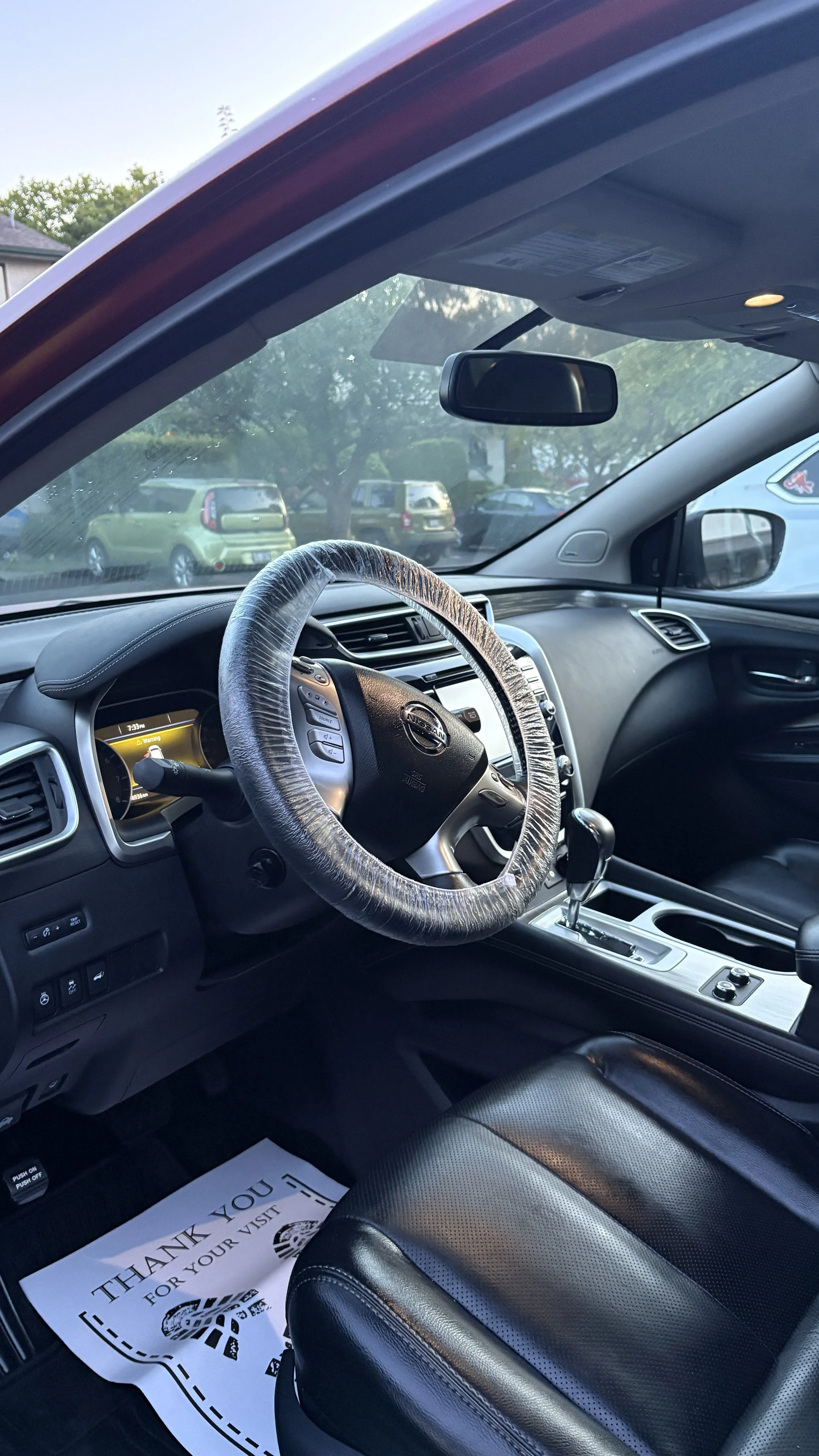 Inside view of a Nissan vehicle with a black leather seat, steering wheel with cover, gear shift, and a 'Thank You' mat on the floor. Seen through the windshield are parked cars and trees outside.