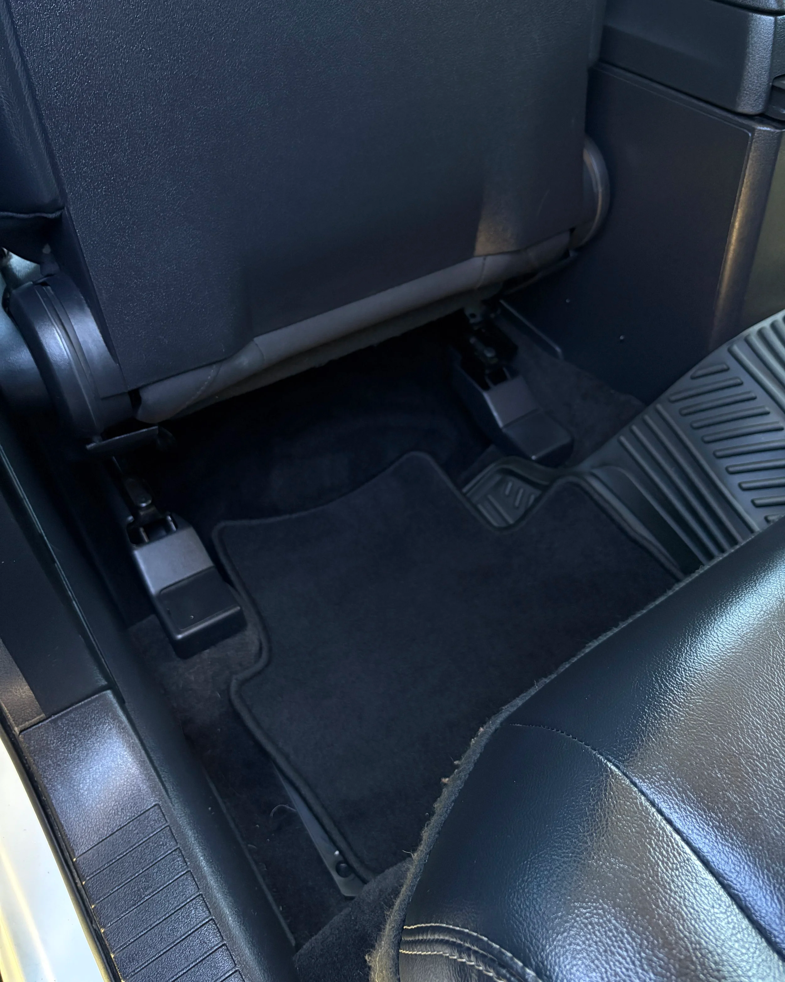 The image shows the interior of a vehicle, specifically the area behind the front seats. Visible elements include the back of the driver or front passenger seat, a plastic cover around the seat's adjustment mechanism, the vehicle's floor mat, and part of the center console.