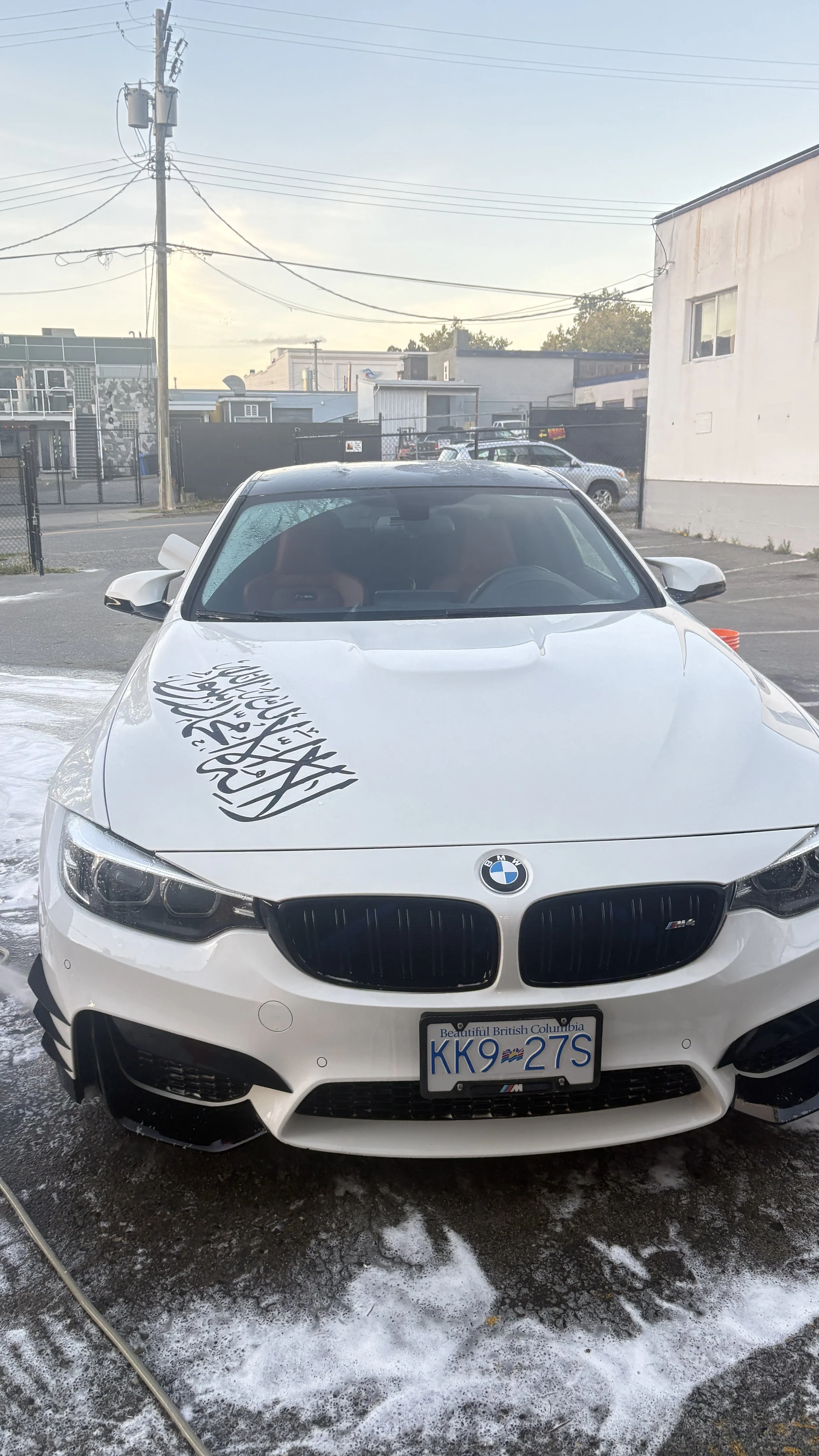 White BMW car with Saudi Arabia flag design on the hood, parked in a wet parking lot with foam and cleaning supplies nearby