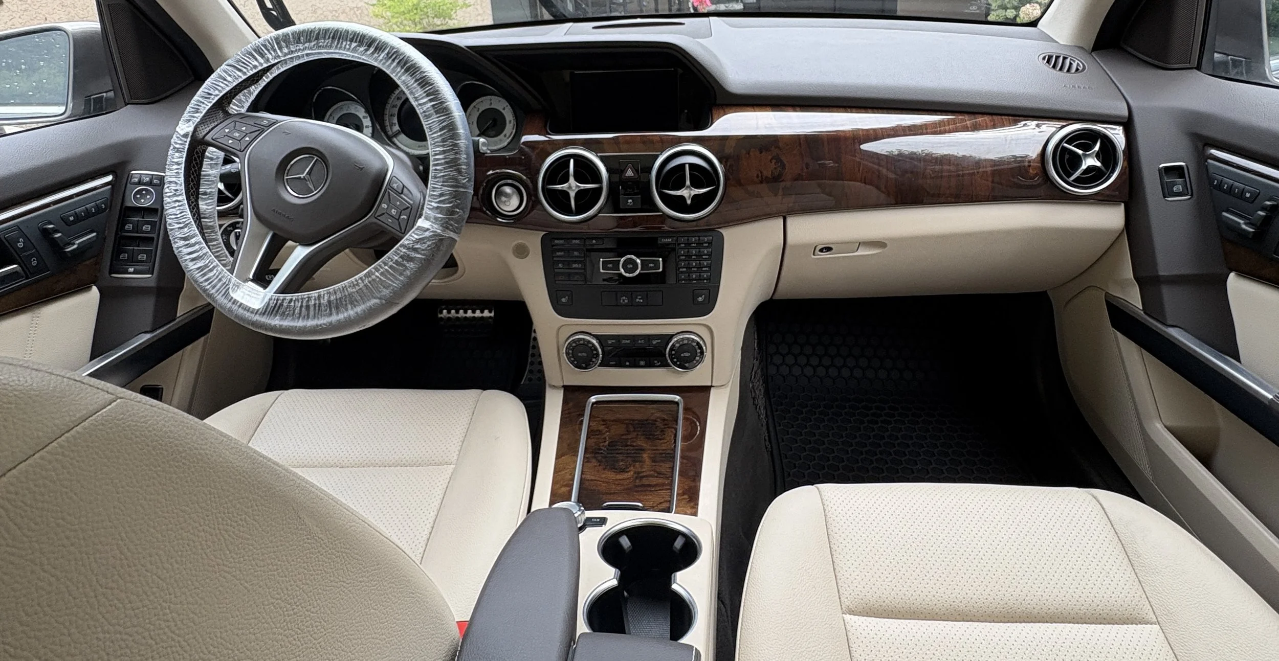 Interior of a luxury car, beige seats, wooden dashboard, steering wheel covered in plastic.