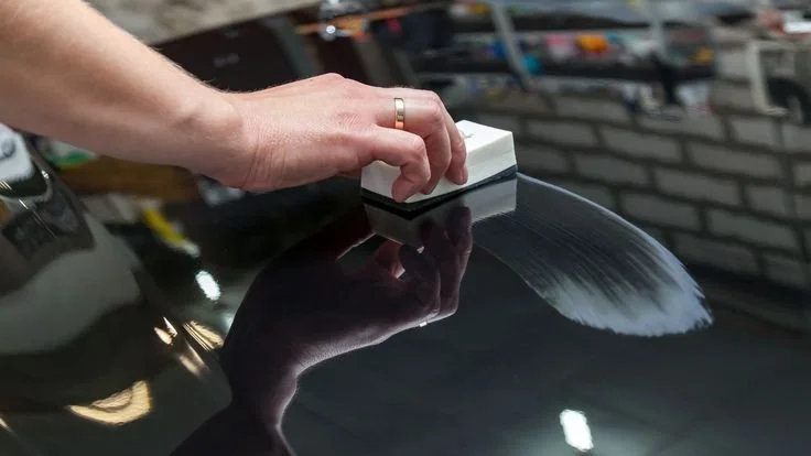 Hand polishing the surface of a black car with a polishing pad in a garage.