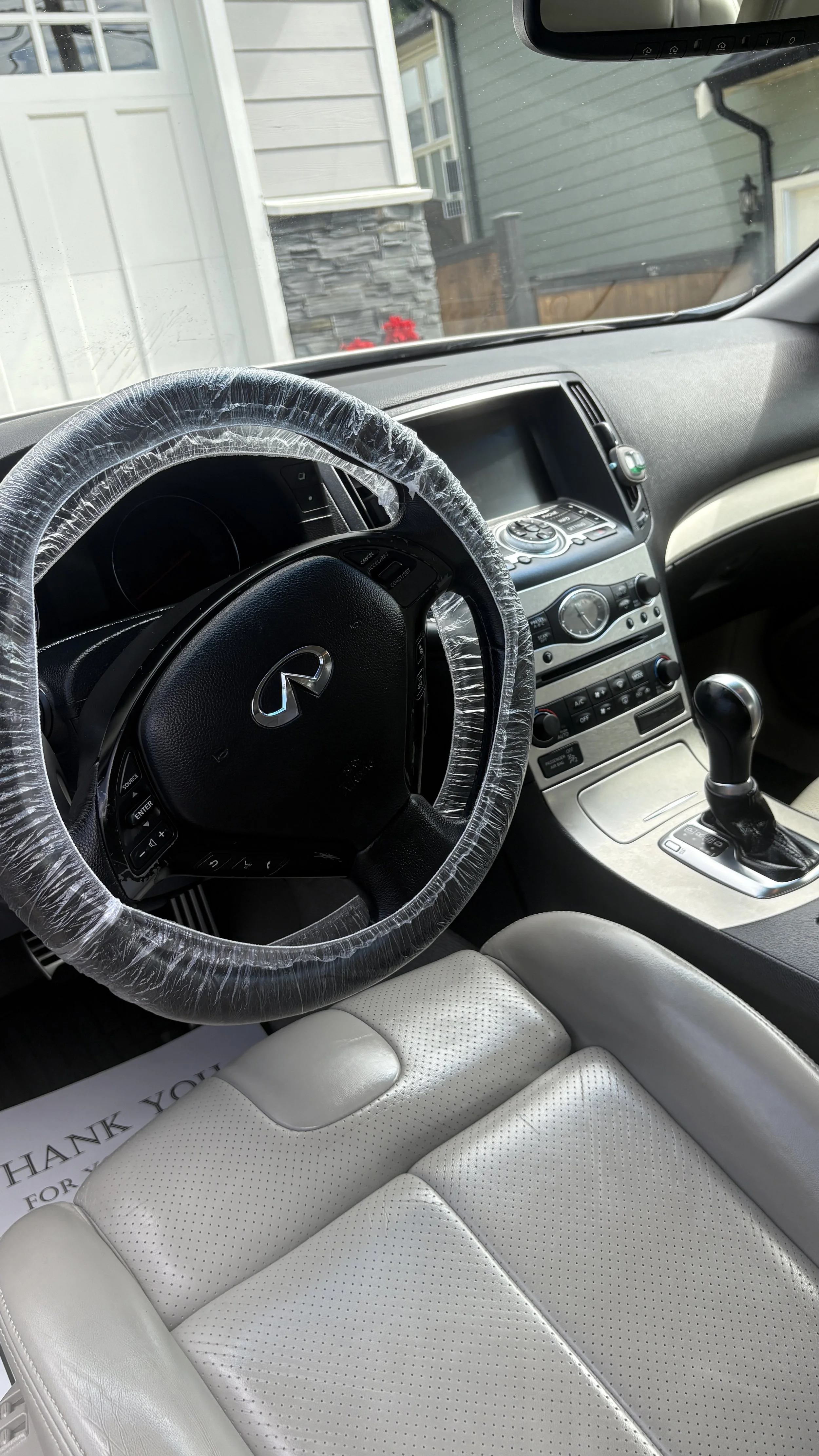 Interior of a Nissan Infiniti car showing the steering wheel with a wrapped cover, gear shift, dashboard, and beige leather seat. Outside through the windshield, part of a house and some red flowers are visible.