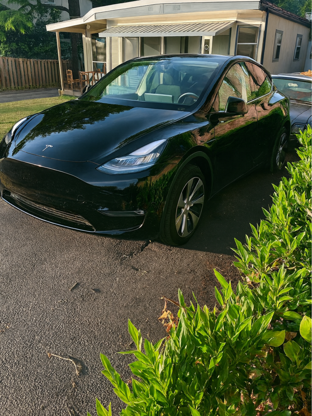 A black Tesla Model 3 parked on a driveway in front of a house with a porch and green bushes.
