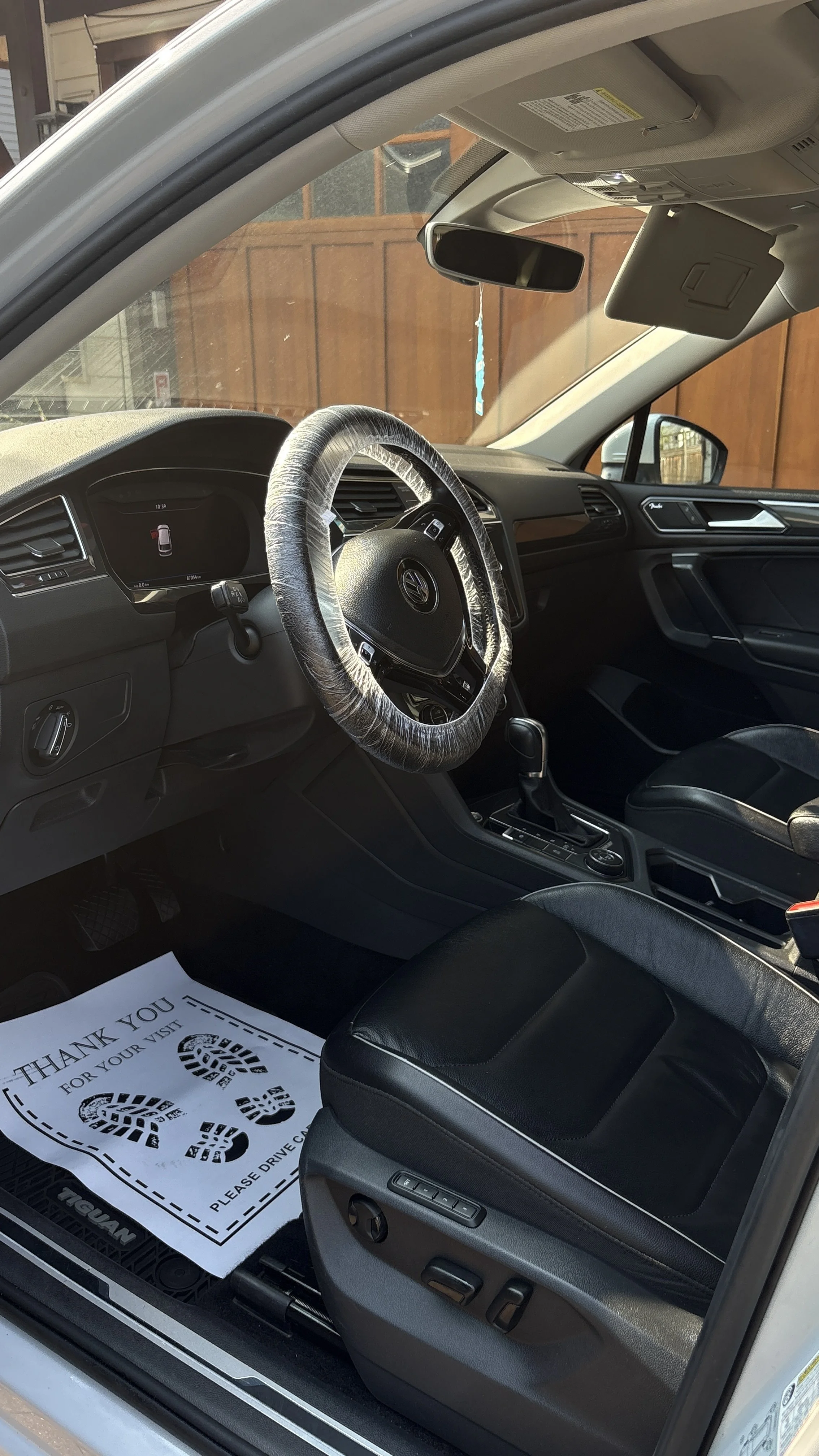 Interior of a Volkswagen car showing the driver seat, steering wheel with a plastic cover, dashboard, rearview mirror, and a paper floor mat with footprints printed on it.