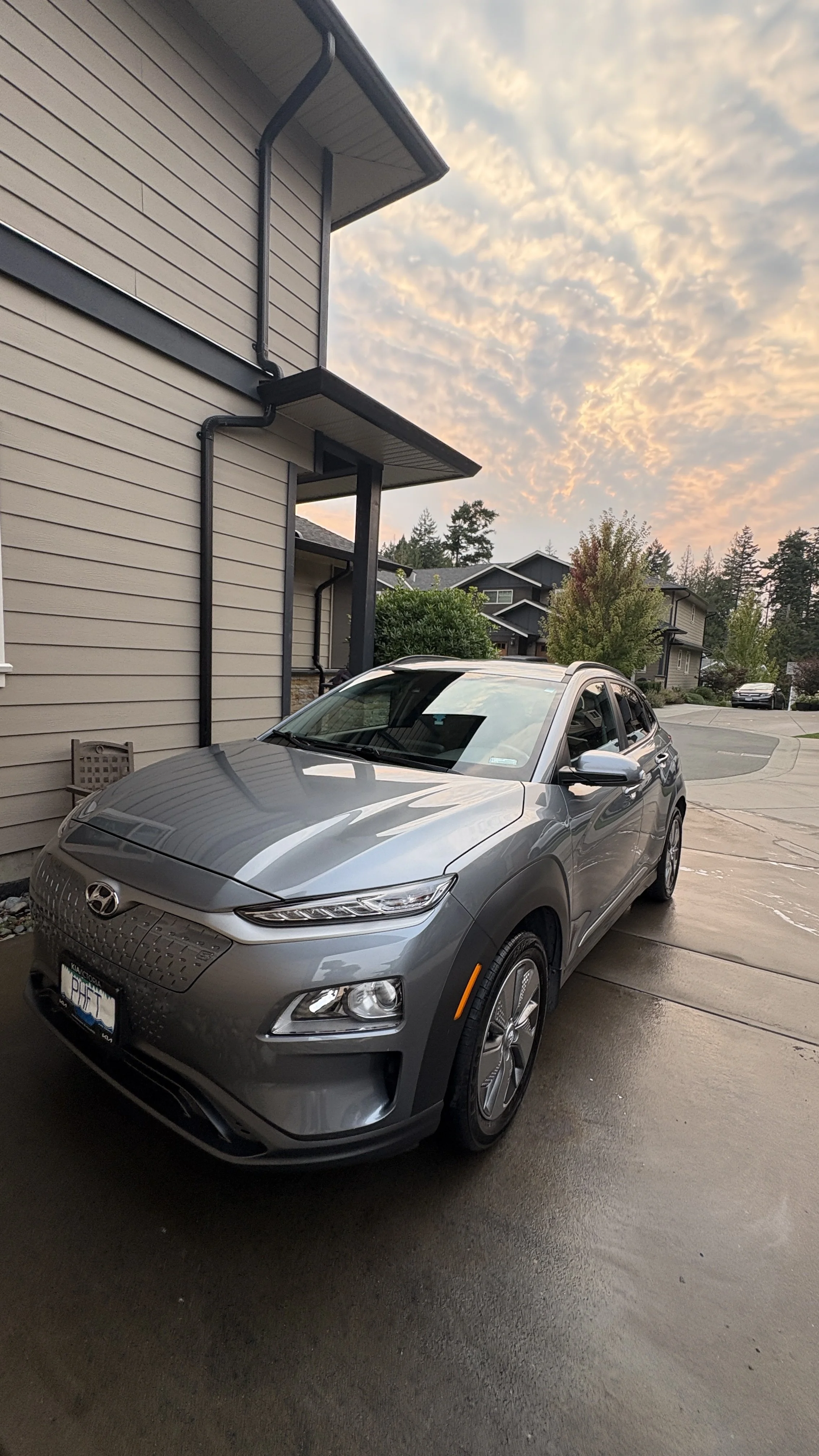 A silver Hyundai Kona parked on a wet driveway in front of a beige house with black gutters, with the sky showing a colorful cloud pattern at sunset.