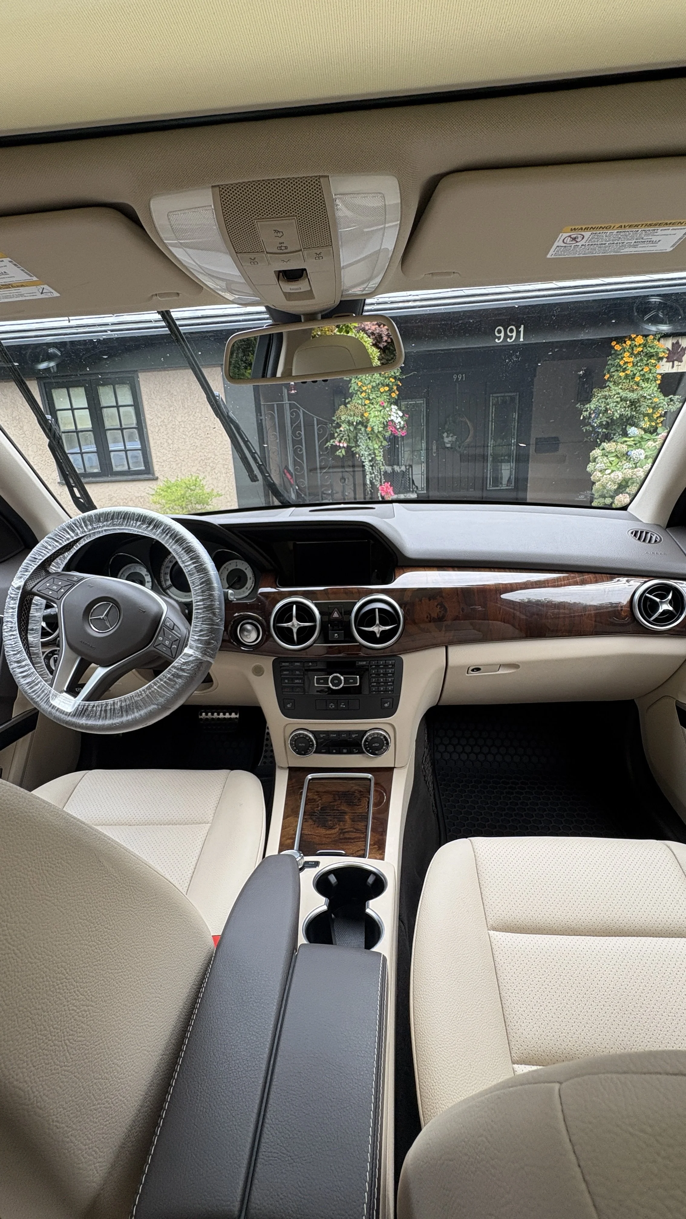 Interior view of a luxury car showing a steering wheel, dashboard with wood accents, beige leather seats, and a rearview mirror, with a house and flowers visible outside through the windshield.