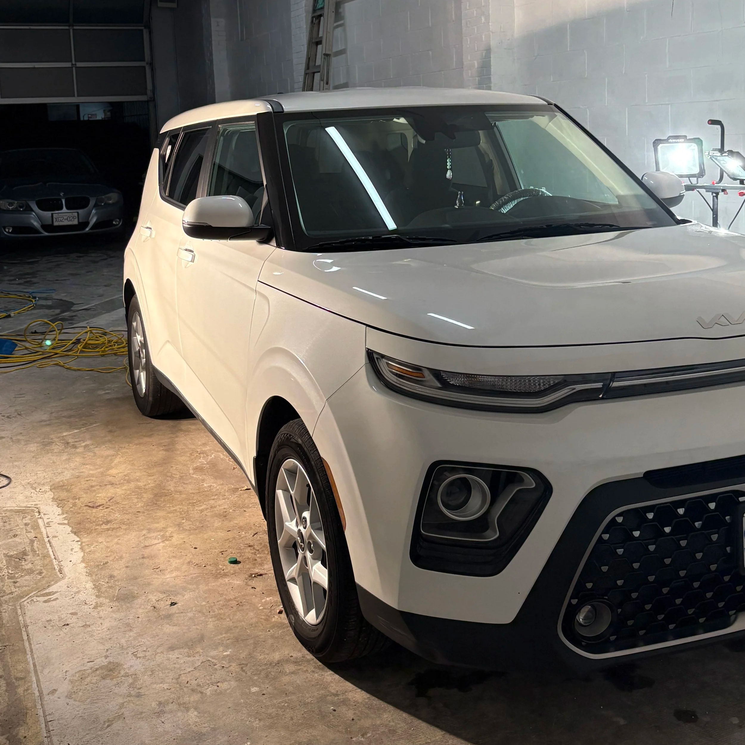 White SUV parked indoors, with a gray car in the background and studio lighting equipment visible.