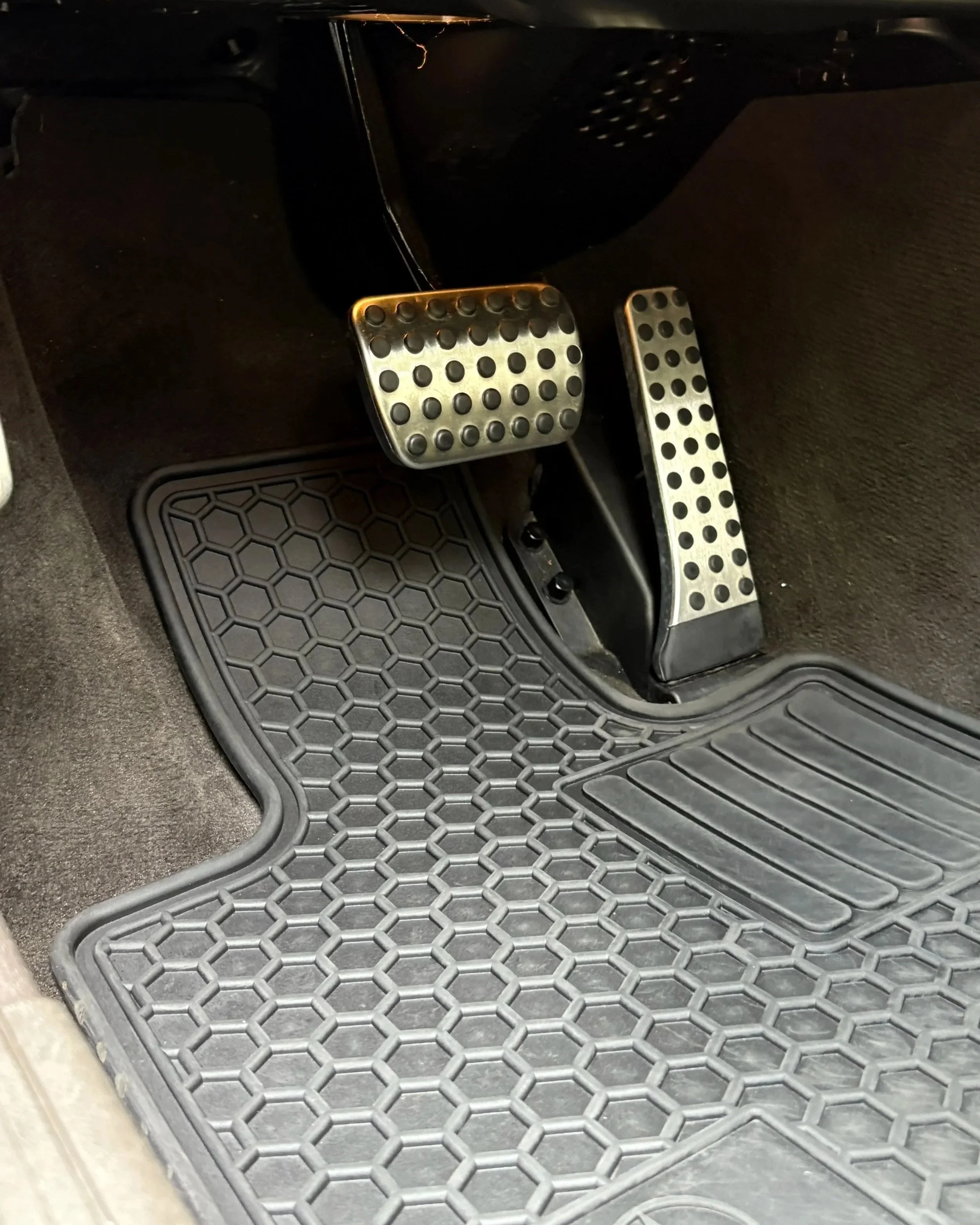 Close-up of a car's foot pedals, including the accelerator, brake, and clutch, with a honeycomb-pattern rubber floor mat underneath.