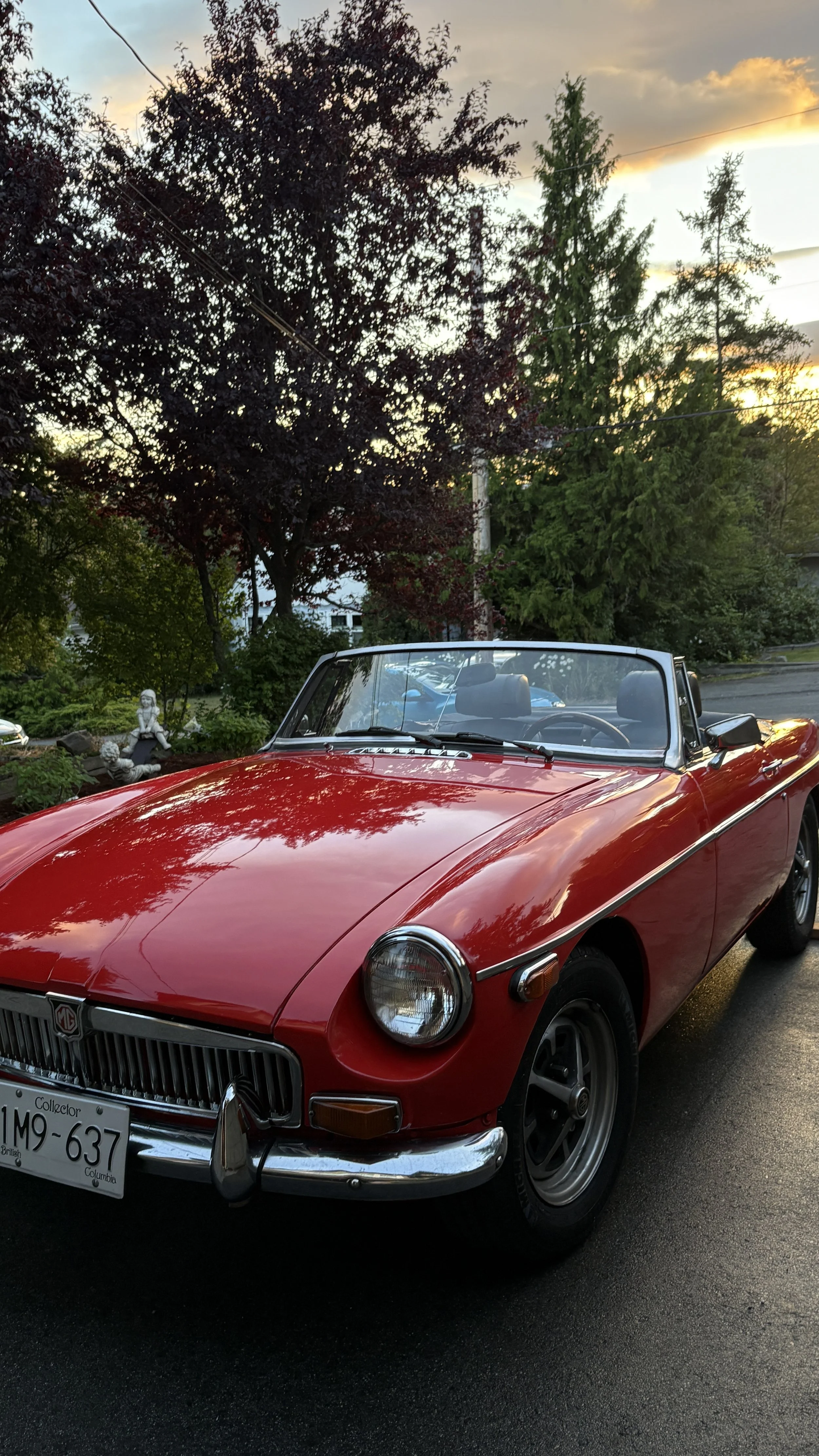 A red vintage MG convertible car parked on a street at sunset, with trees and a garden in the background.