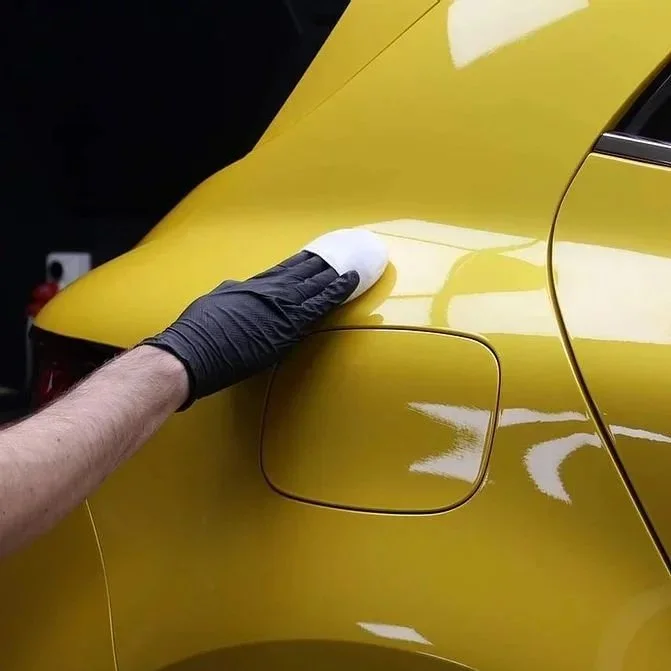 Person wearing a black glove cleaning the yellow car's rear fender with a white cloth.