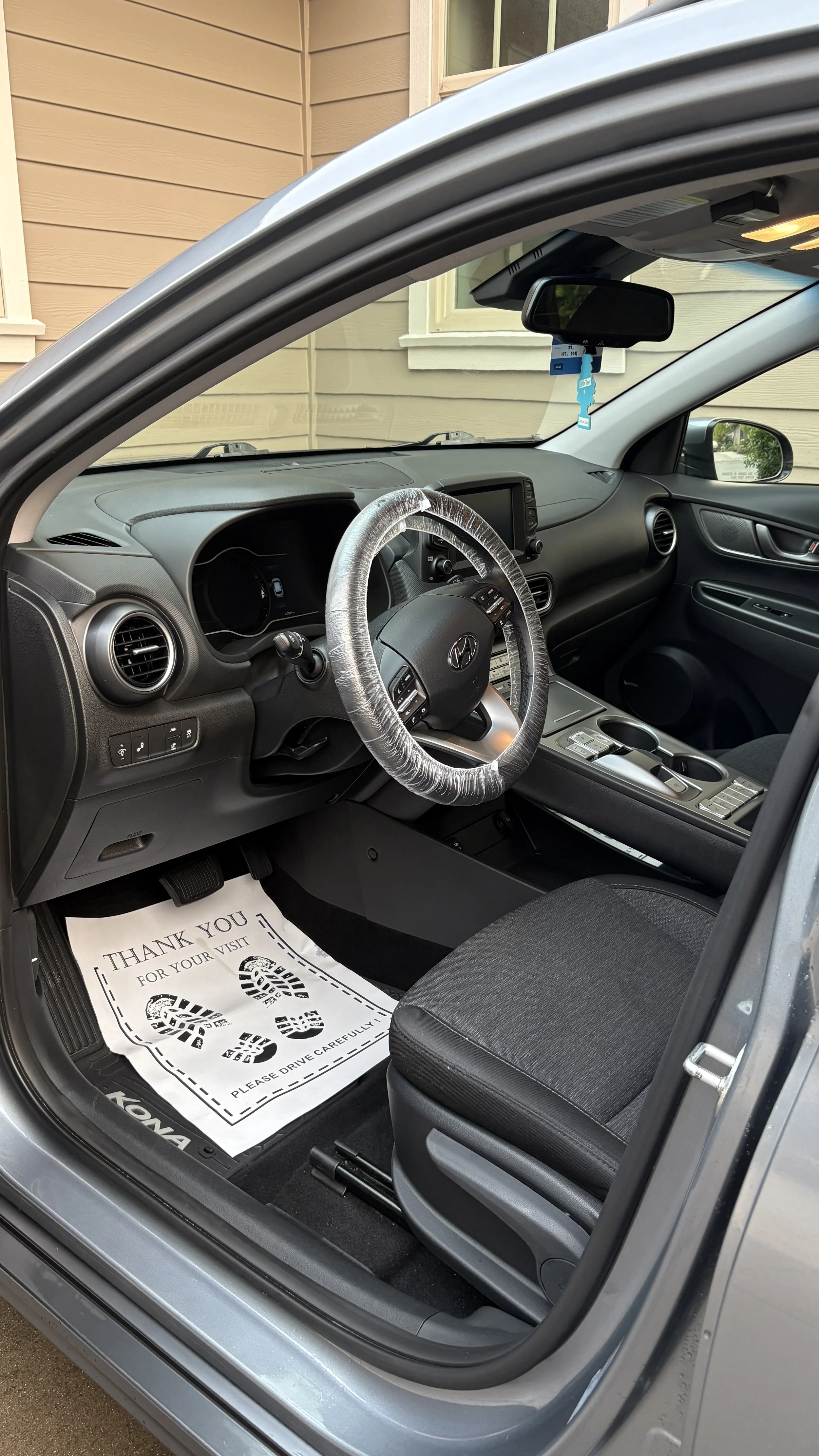 Interior of a Hyundai Kona, showing the driver's seat, steering wheel with a plastic cover, dashboard, and center console. A paper floor mat reads 'Thank you for your visit' and 'Please drive carefully.'