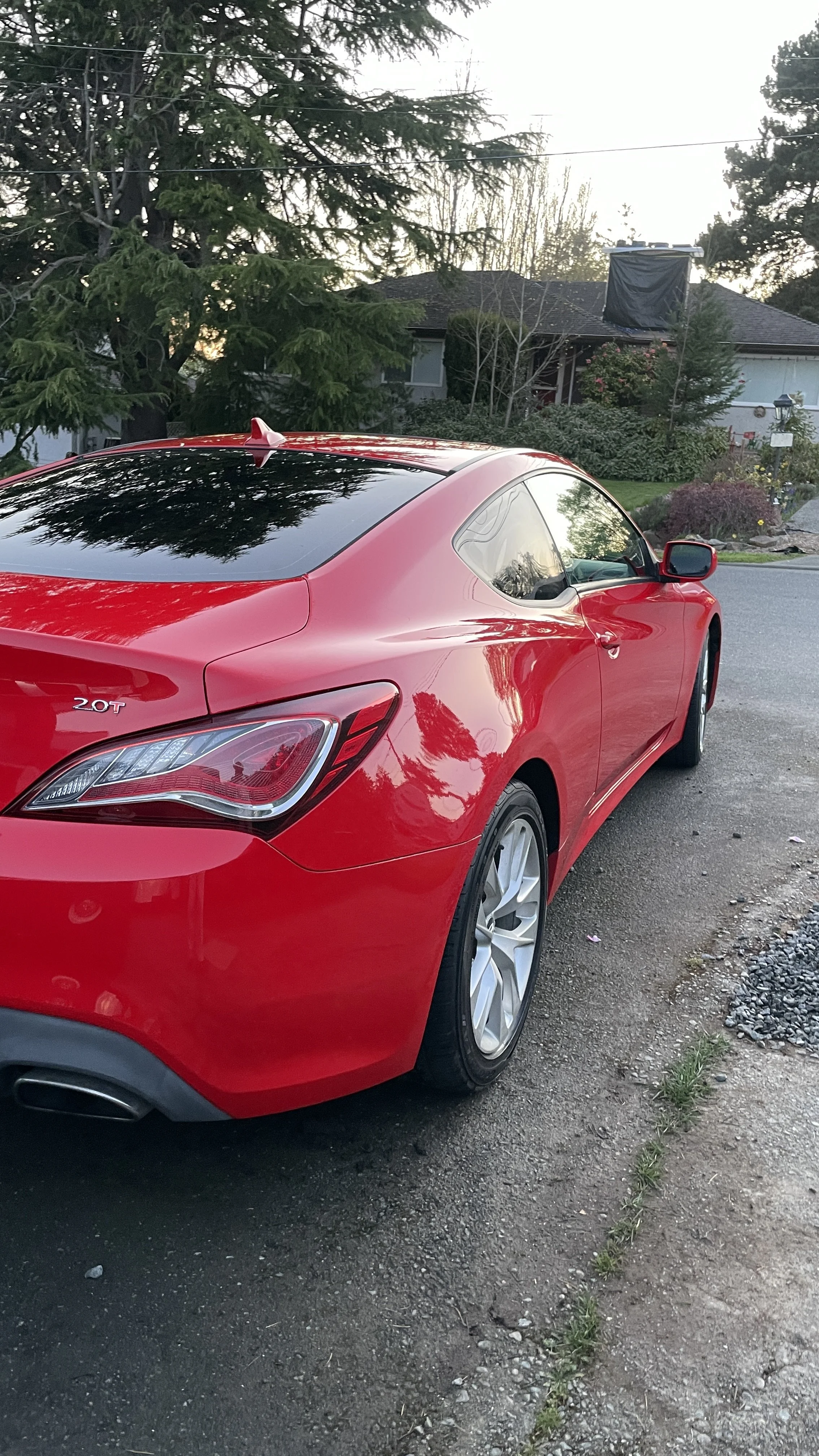 Red coupe car parked on a driveway with trees and houses in the background during twilight.