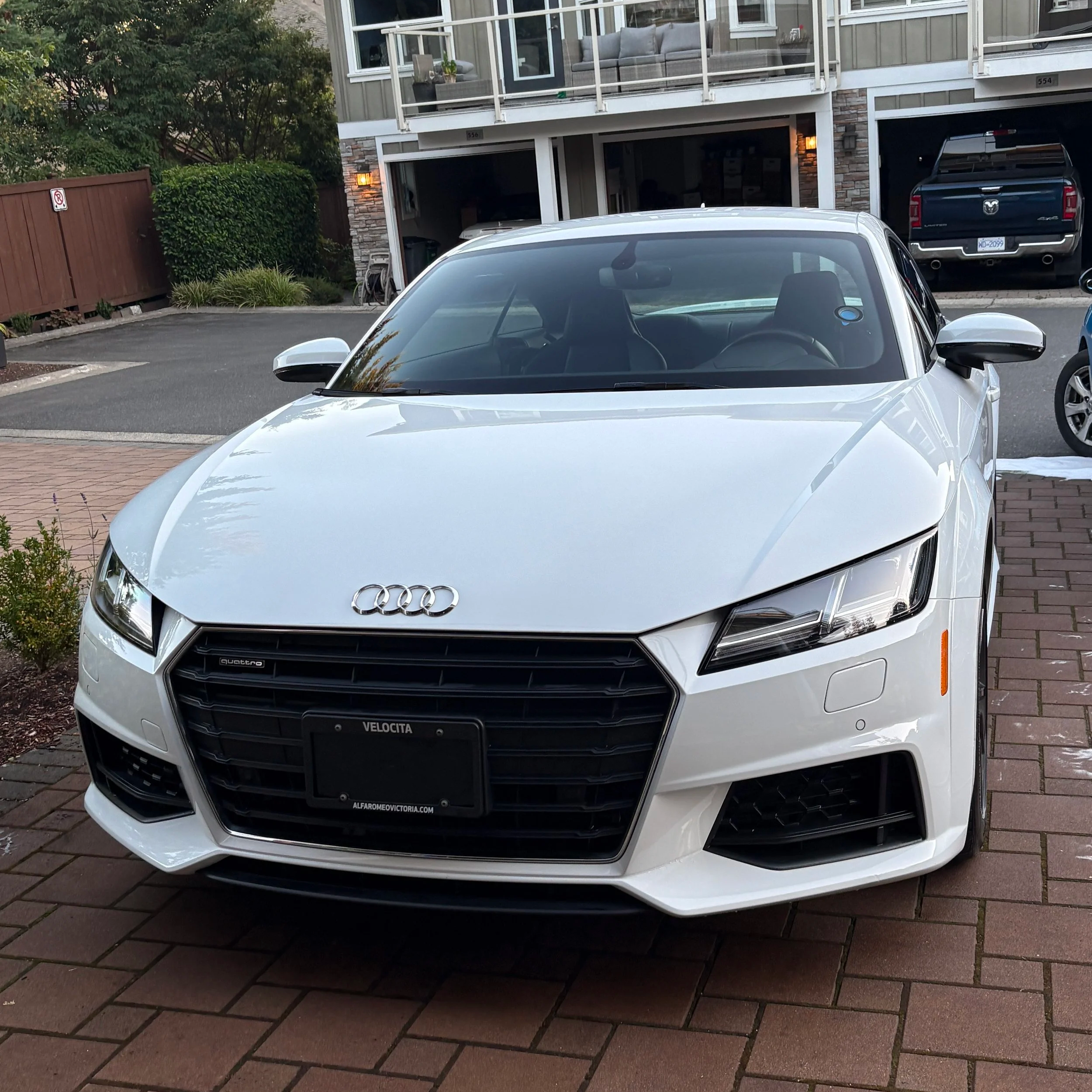 A white Audi car parked in a residential driveway with garage and apartment building in the background.