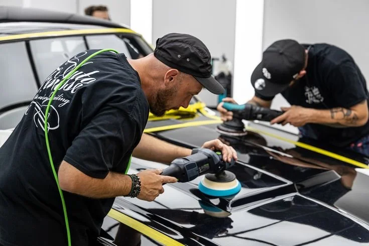 Two men polishing a black car with handheld buffers at a workshop.