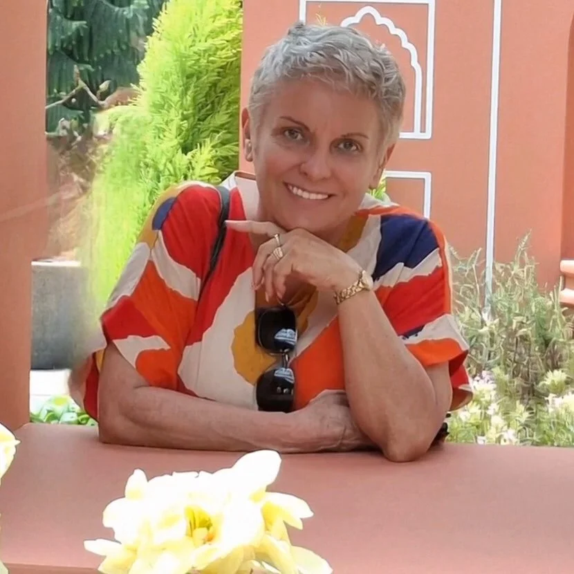 A smiling woman with short gray hair sitting at a table indoors, wearing a colorful patterned shirt and a watch. She has sunglasses hanging from her shirt and is resting her chin on her hand. There are yellow flowers on the table and a garden visible through the open window behind her.