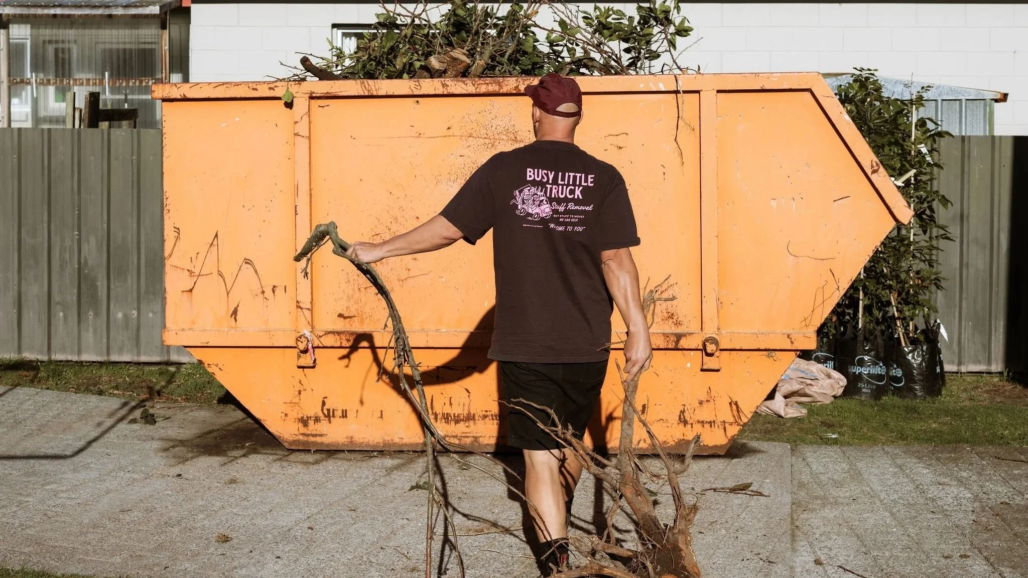 A person, wearing a black t-shirt with the words 'BUSY LITTLE TRUCK' on the back, is using a garden tool to clear branches on a pile of yard debris in front of a large orange dumpster.